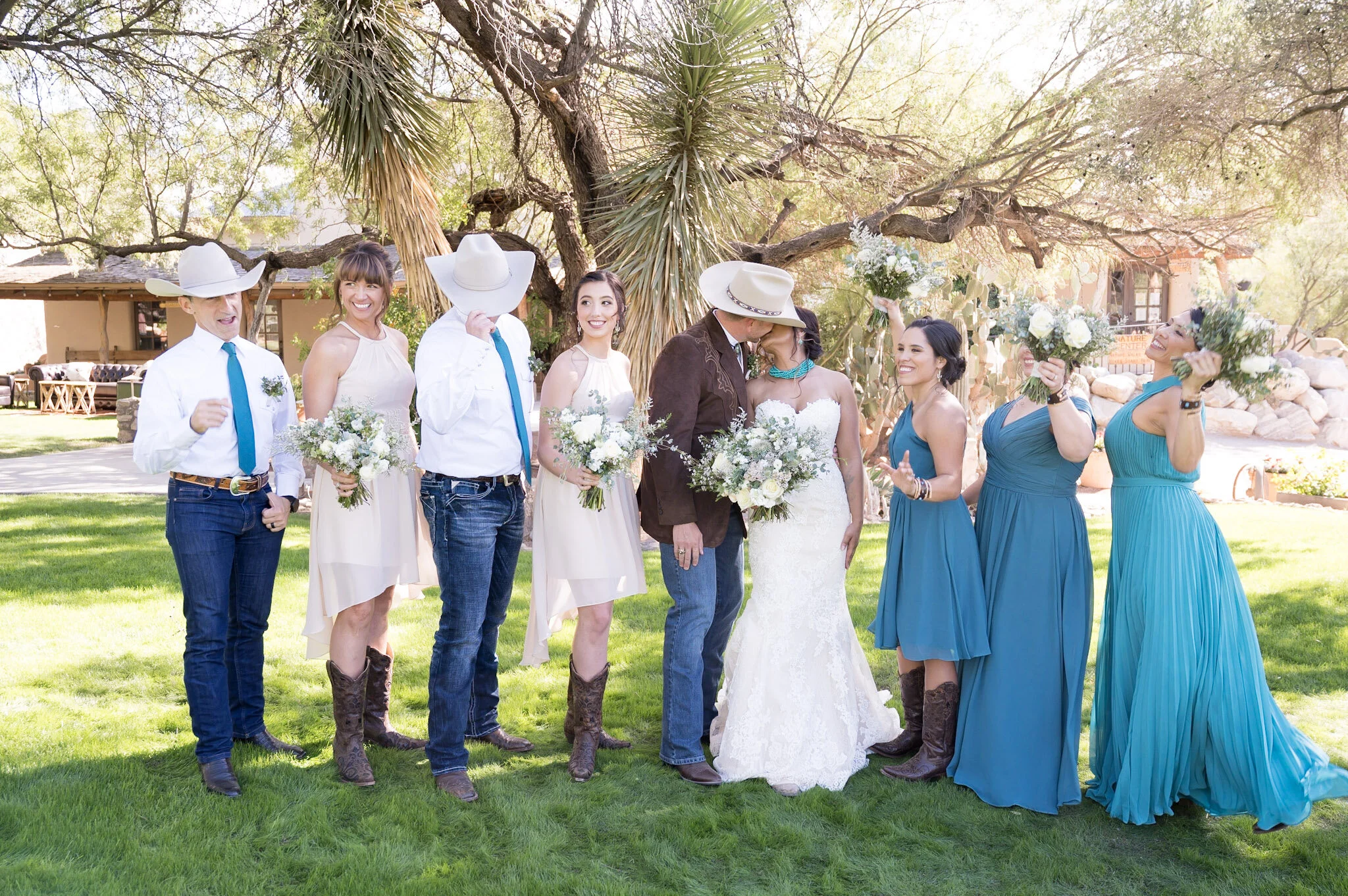 A group of people at a wedding, with some dressed as cowboys and cowgirls, standing outdoors on a green lawn under trees, with a couple in wedding attire in the center, holding bouquets, embracing and kissing.