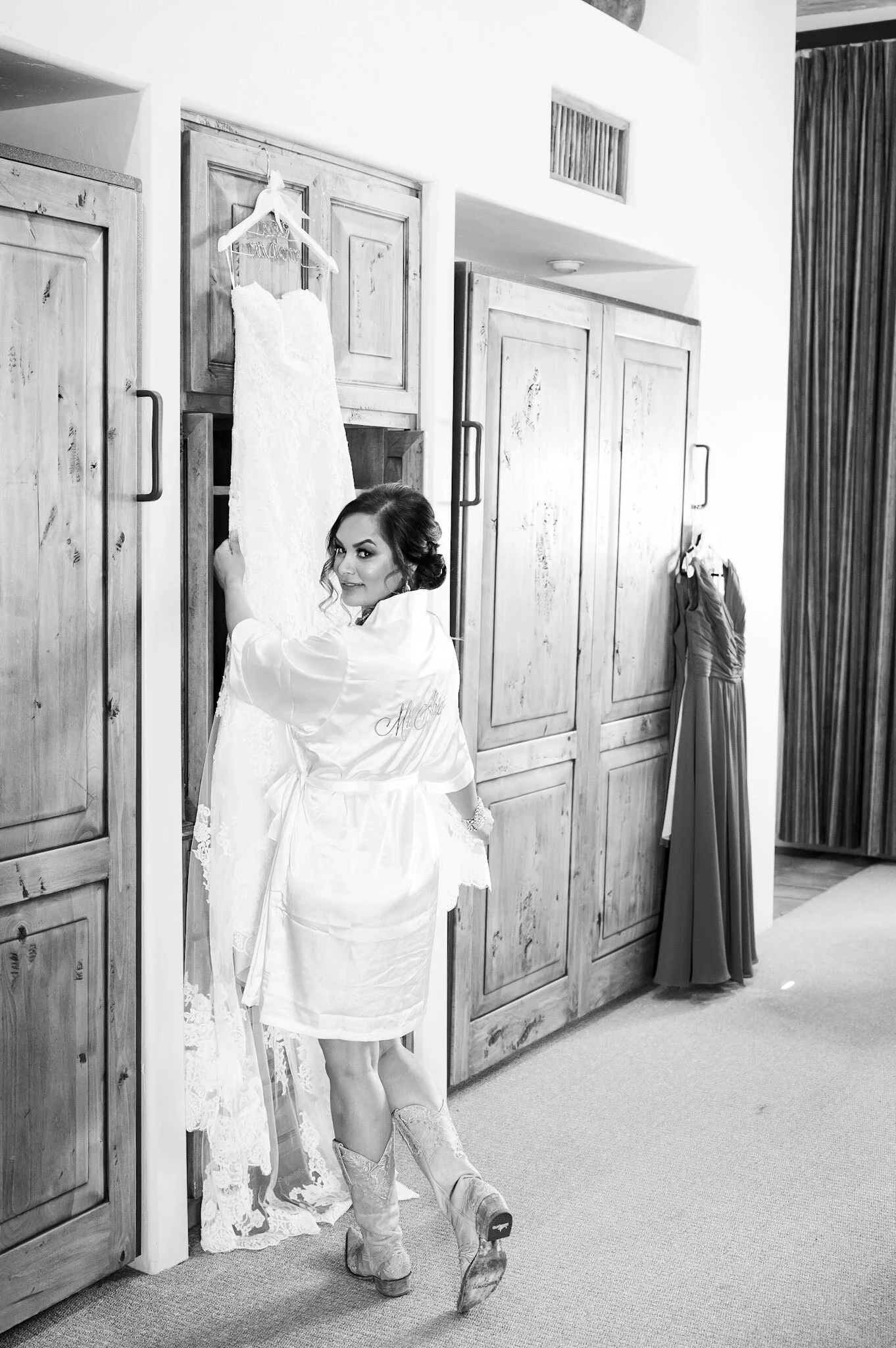 A woman in a white robe is standing in front of wooden cabinet doors, looking over her shoulder and smiling. A wedding dress hangs on the cabinet. There are curtains and another dress on a hanger in the background.