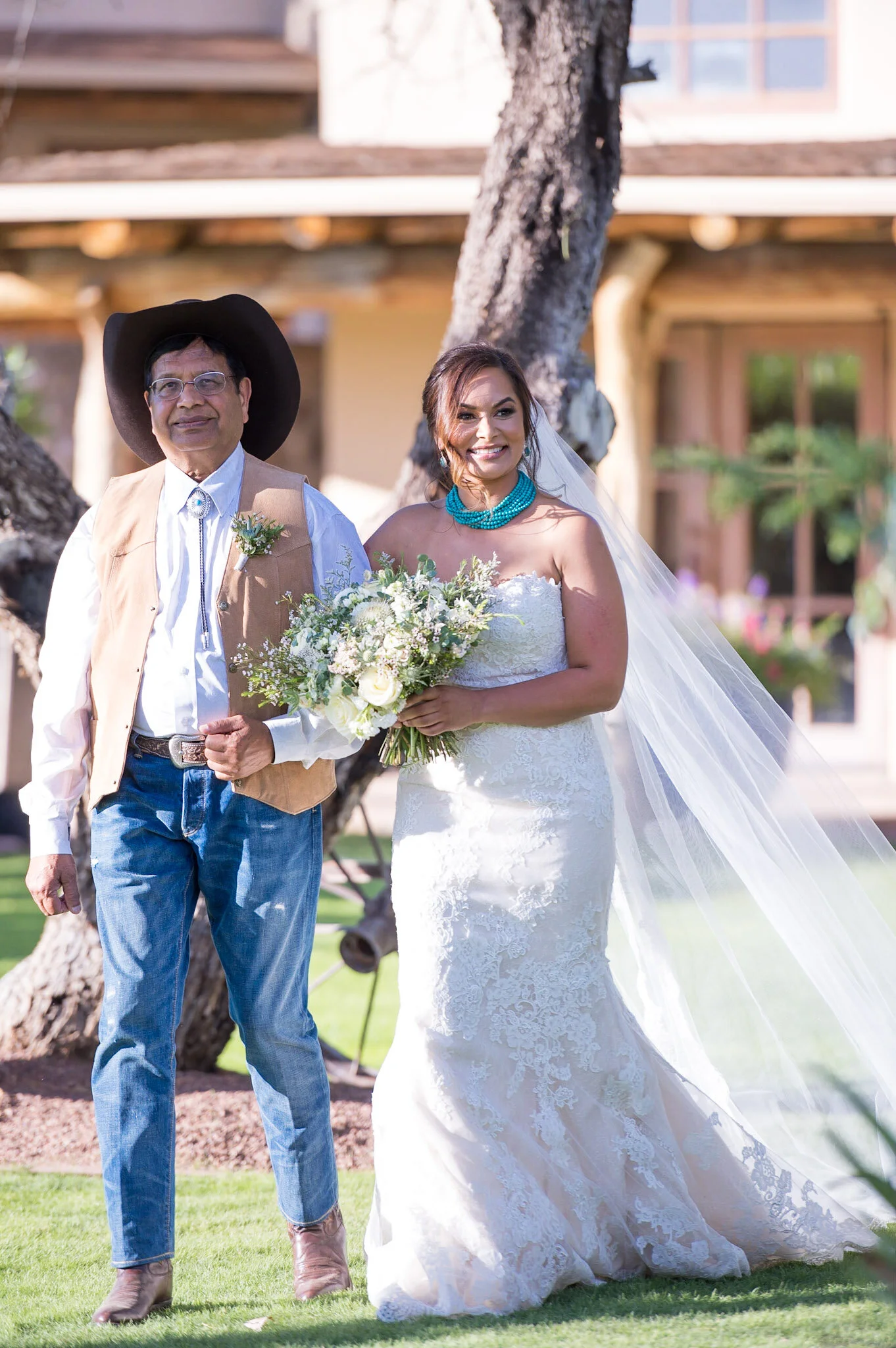 A woman in a white wedding dress holding a bouquet of flowers, standing next to a man in western attire, outdoors, with trees and a building in the background.