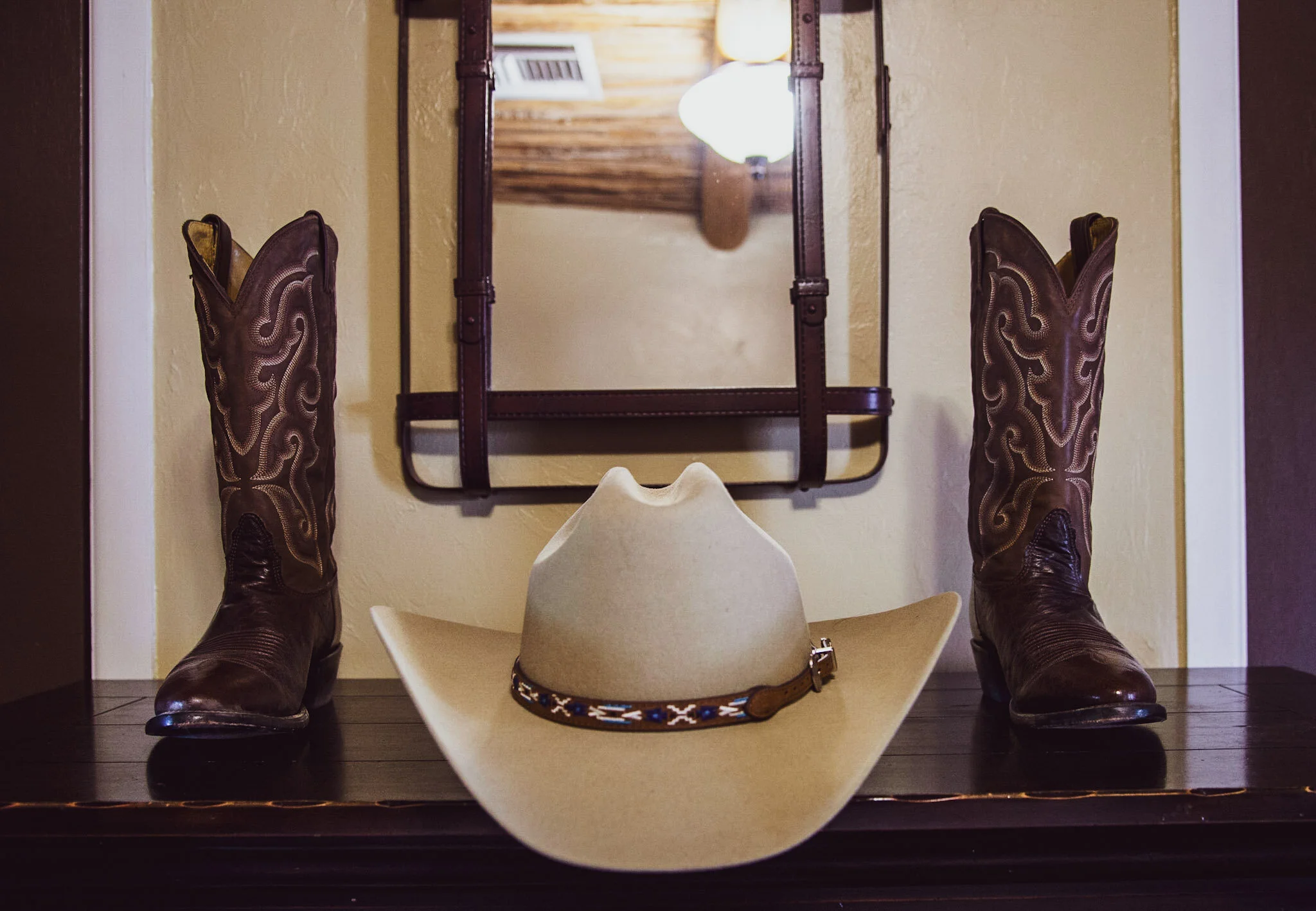 A person wearing cowboy boots and a cowboy hat, standing in front of a mirror with a cowboy hat resting on a table.