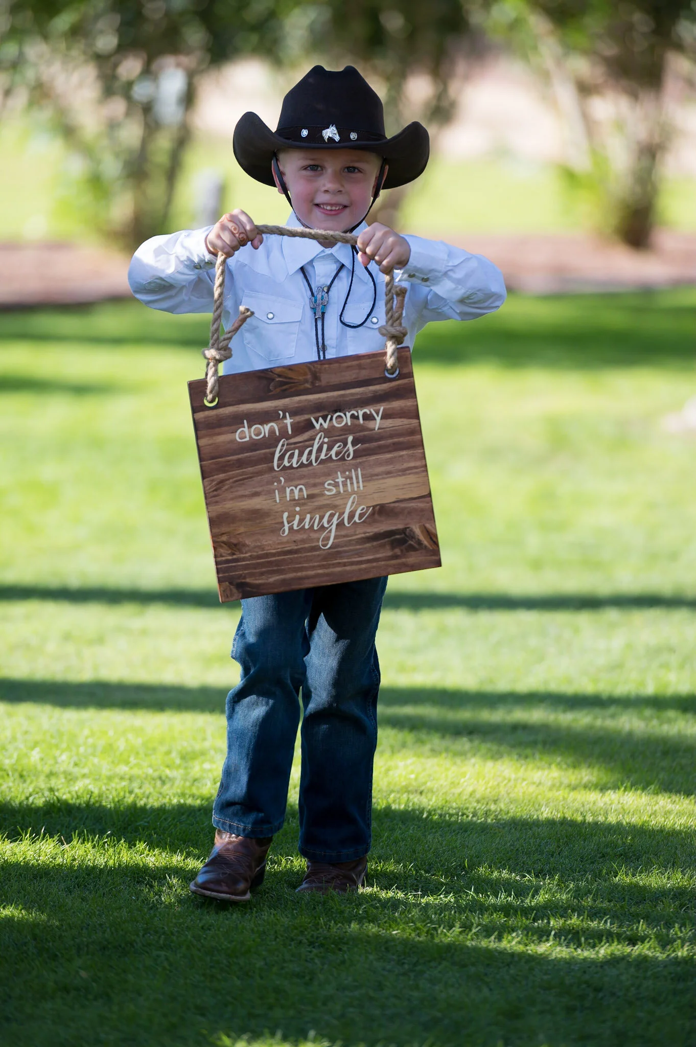 A young boy dressed as a cowboy, wearing a black cowboy hat and blue jeans, standing outdoors on grass and holding a wooden sign that says 'don't worry ladies I'm still single'.