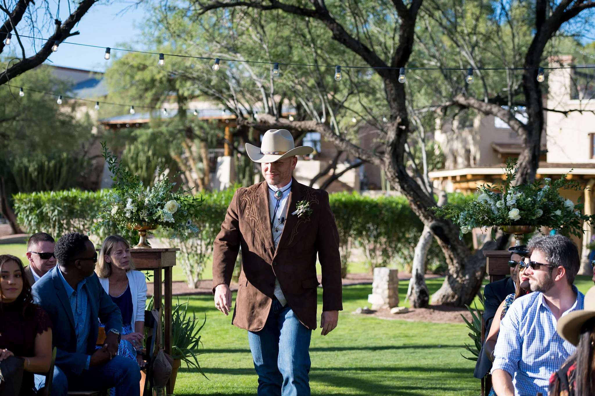 A man wearing a white cowboy hat, brown jacket, and bolo tie standing outdoors among seated people at an event, with string lights and trees in the background.