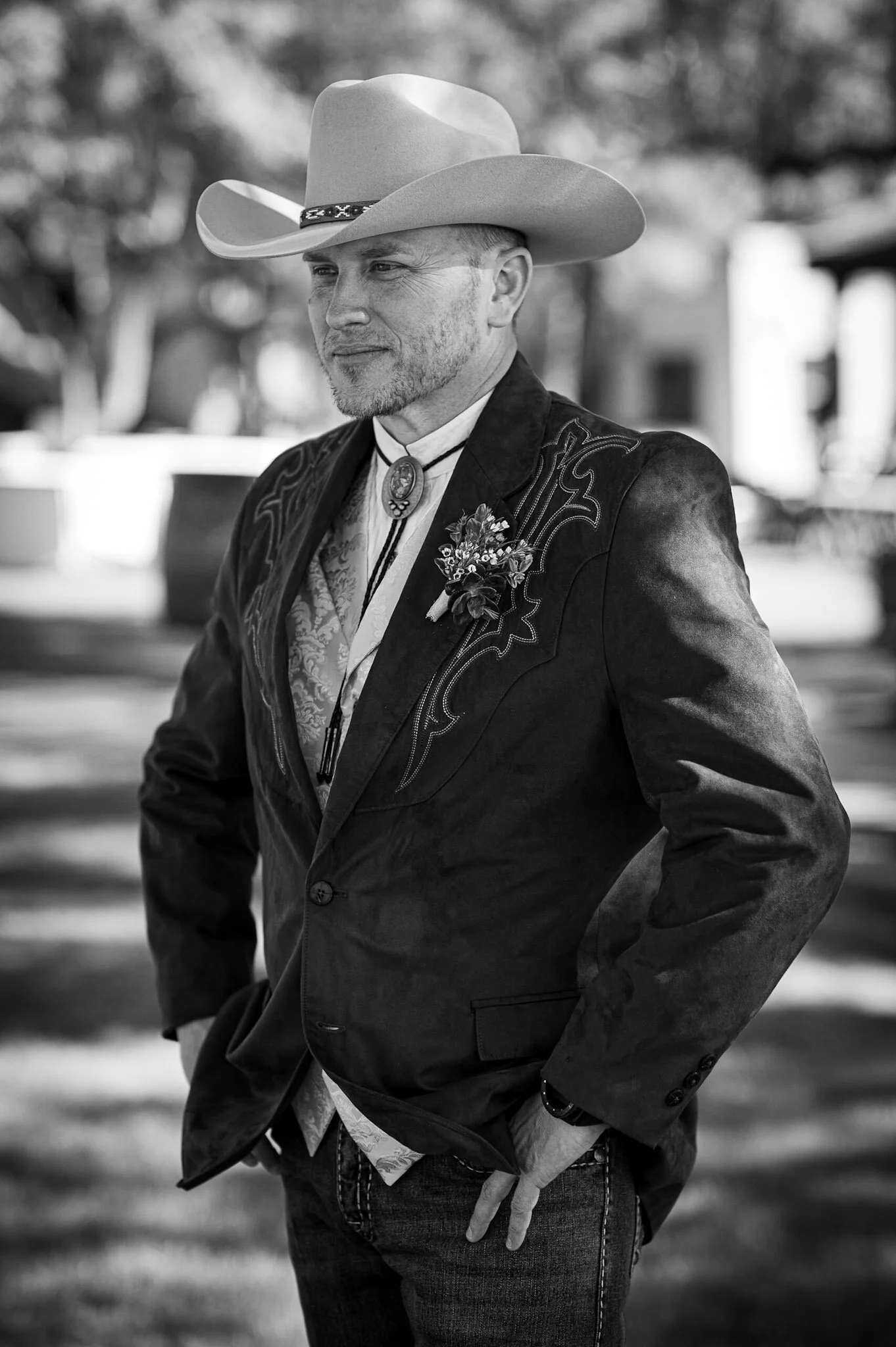 A man dressed in a Western-style outfit, including a cowboy hat, bolo tie, and embroidered jacket, standing outdoors in a casual pose.