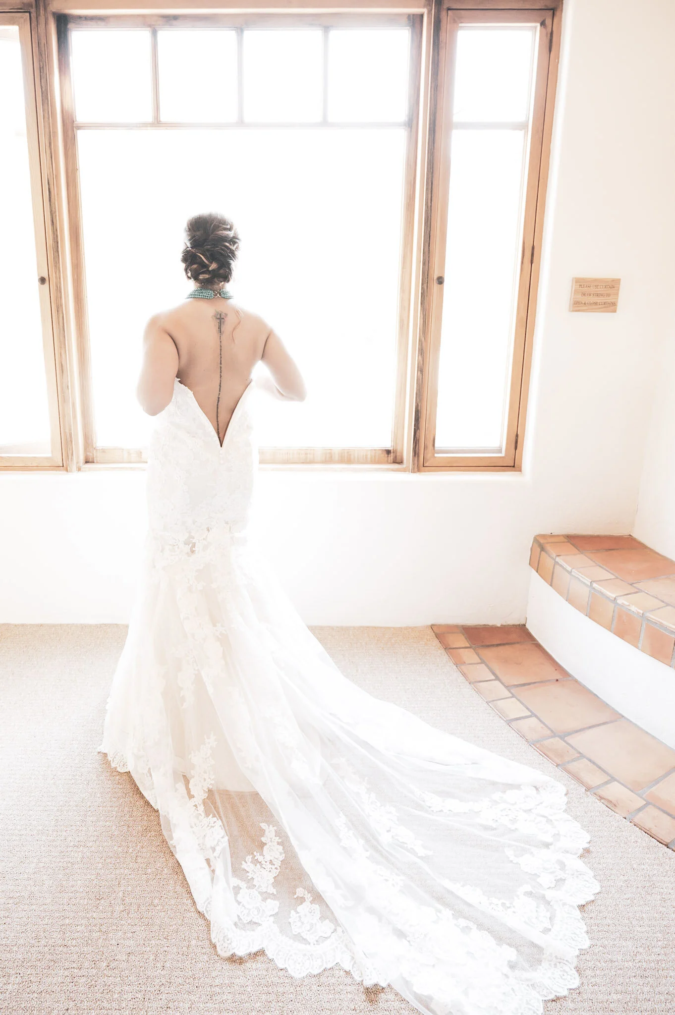 Bride in a white lace wedding dress with a long train standing in front of a window, facing away from the camera.