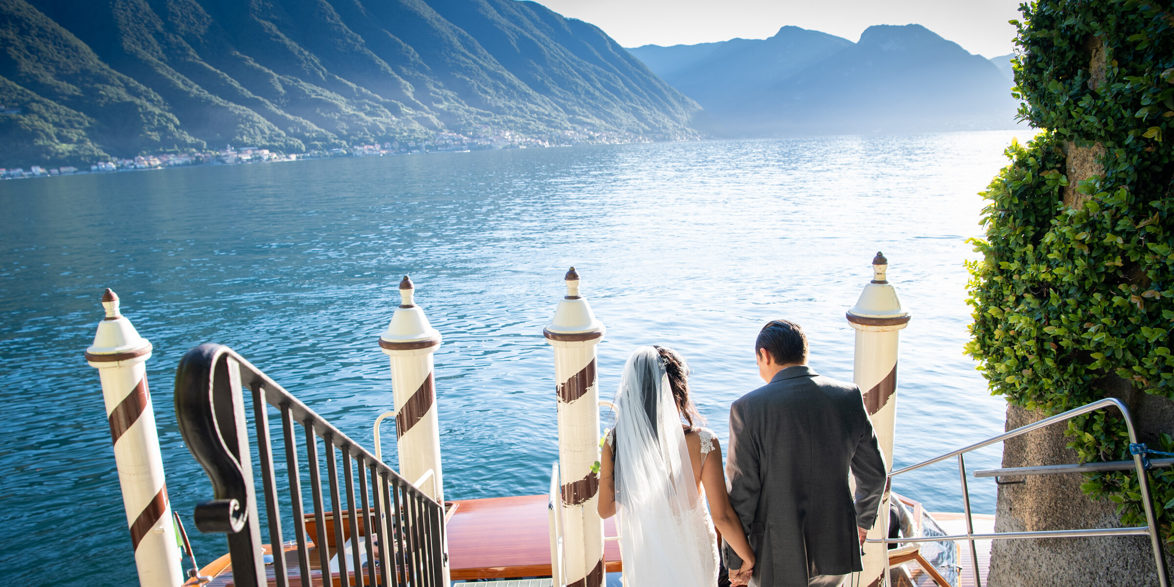 A bride and groom sitting on a boat dock, overlooking a large body of water with mountains in the background and greenery on the side.
