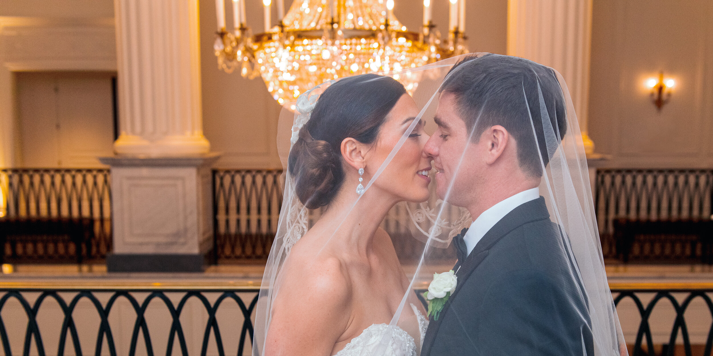 A bride and groom sharing a close, intimate moment during their wedding ceremony, with a chandelier and elegant decor in the background.