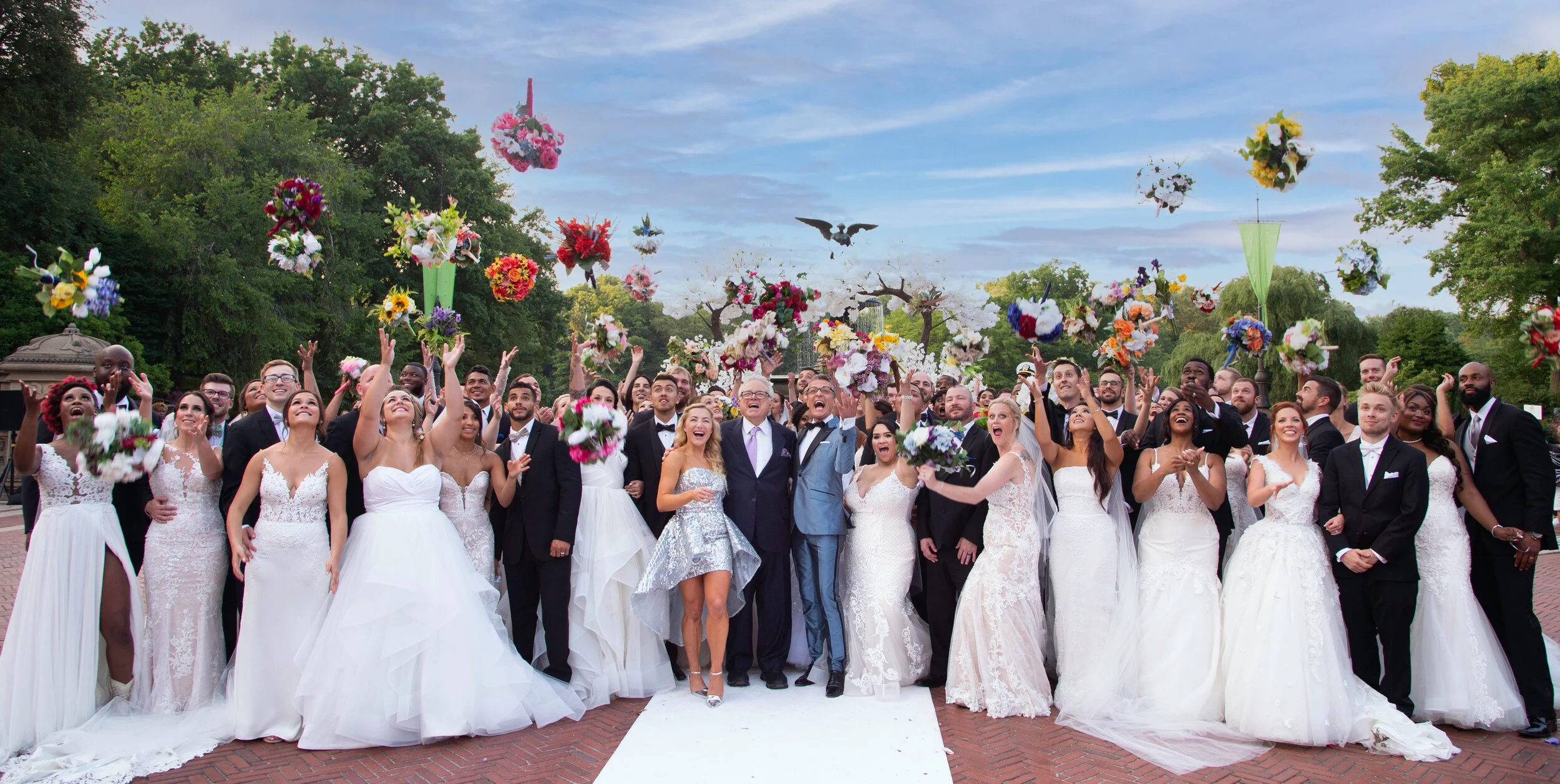 Large group of wedding guests, including brides in white dresses and grooms in suits, celebrating outdoors as flowers are tossed into the air.