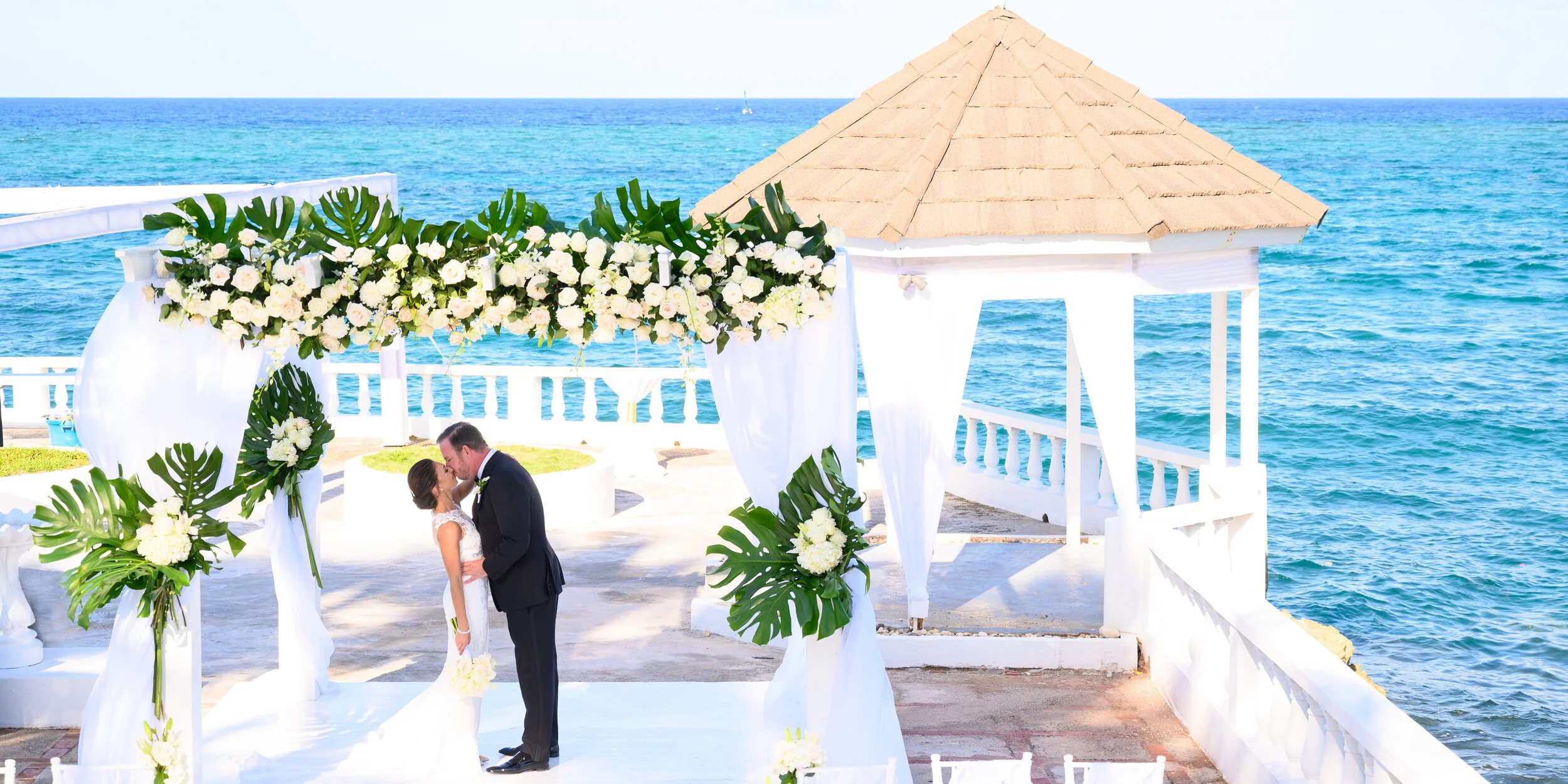 A wedding ceremony taking place outdoors by the sea under a decorated white gazebo with floral arrangements, with a couple kissing.