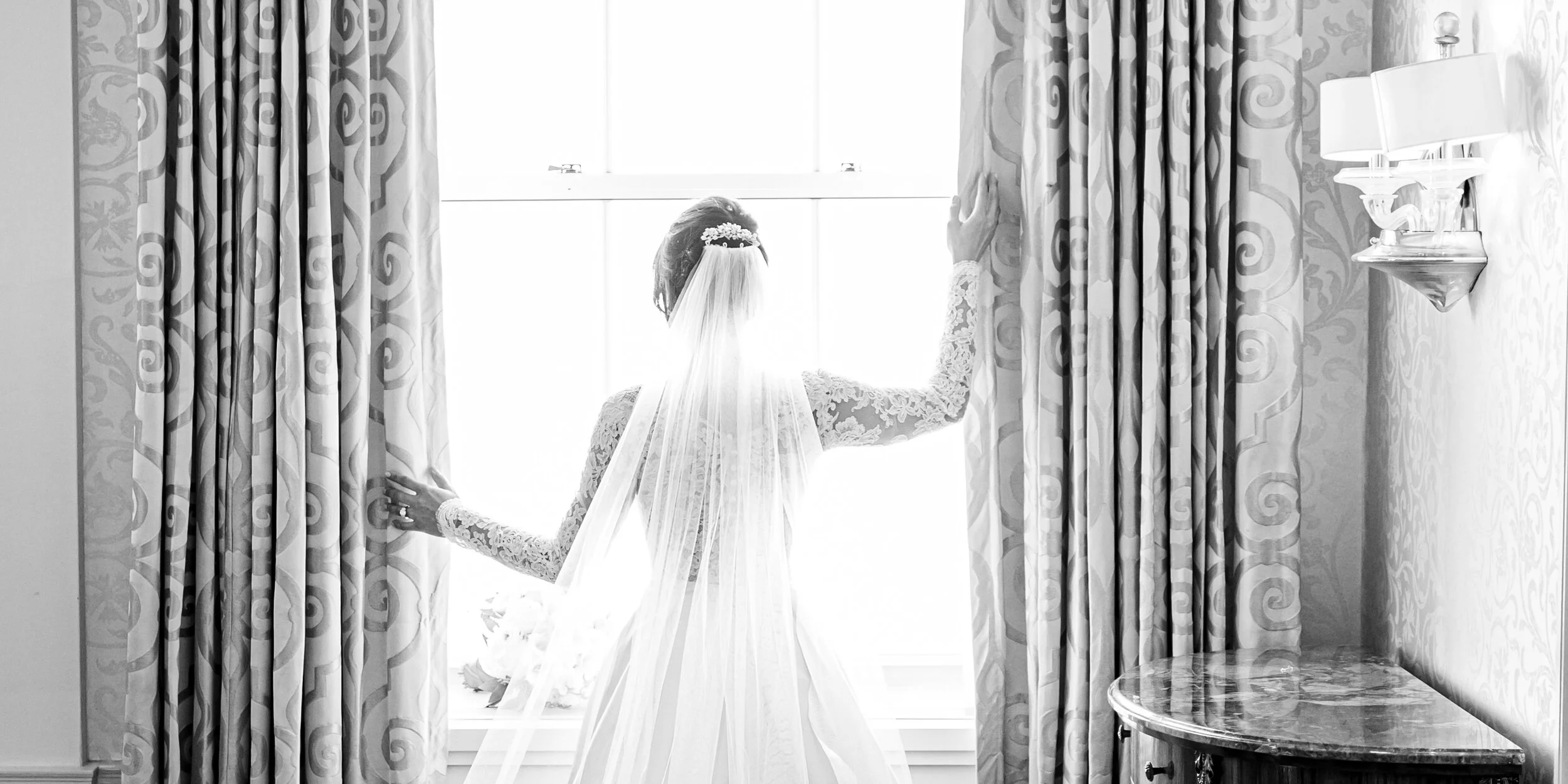 Bride in lace wedding dress and veil looking out a bright window, holding the curtain, with patterned curtains on either side.