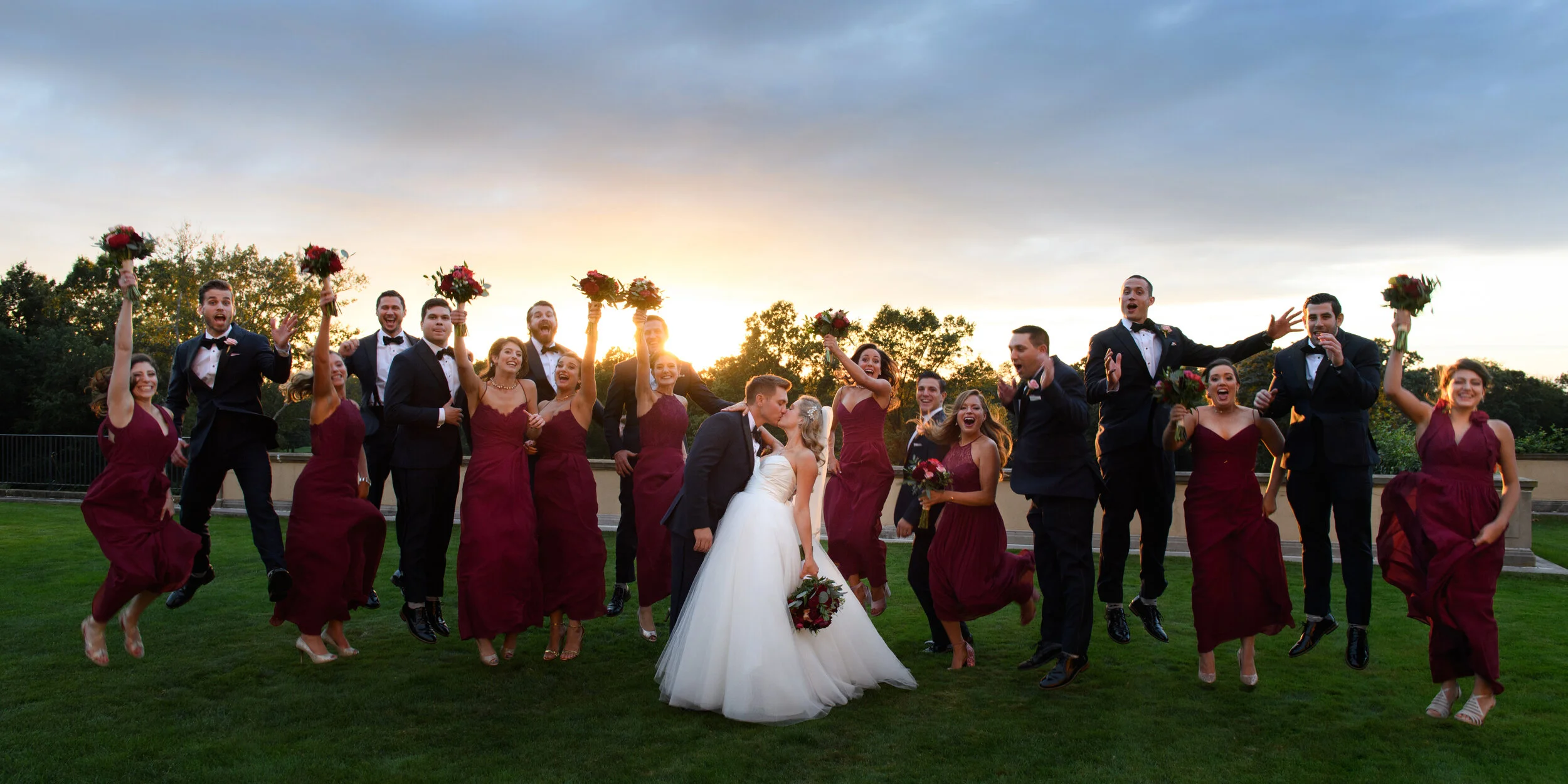 Happy wedding party outside at sunset, with Bride and Groom in the center sharing a kiss surrounded by bridesmaids and groomsmen in formal attire, some holding bouquets and jumping in the air.