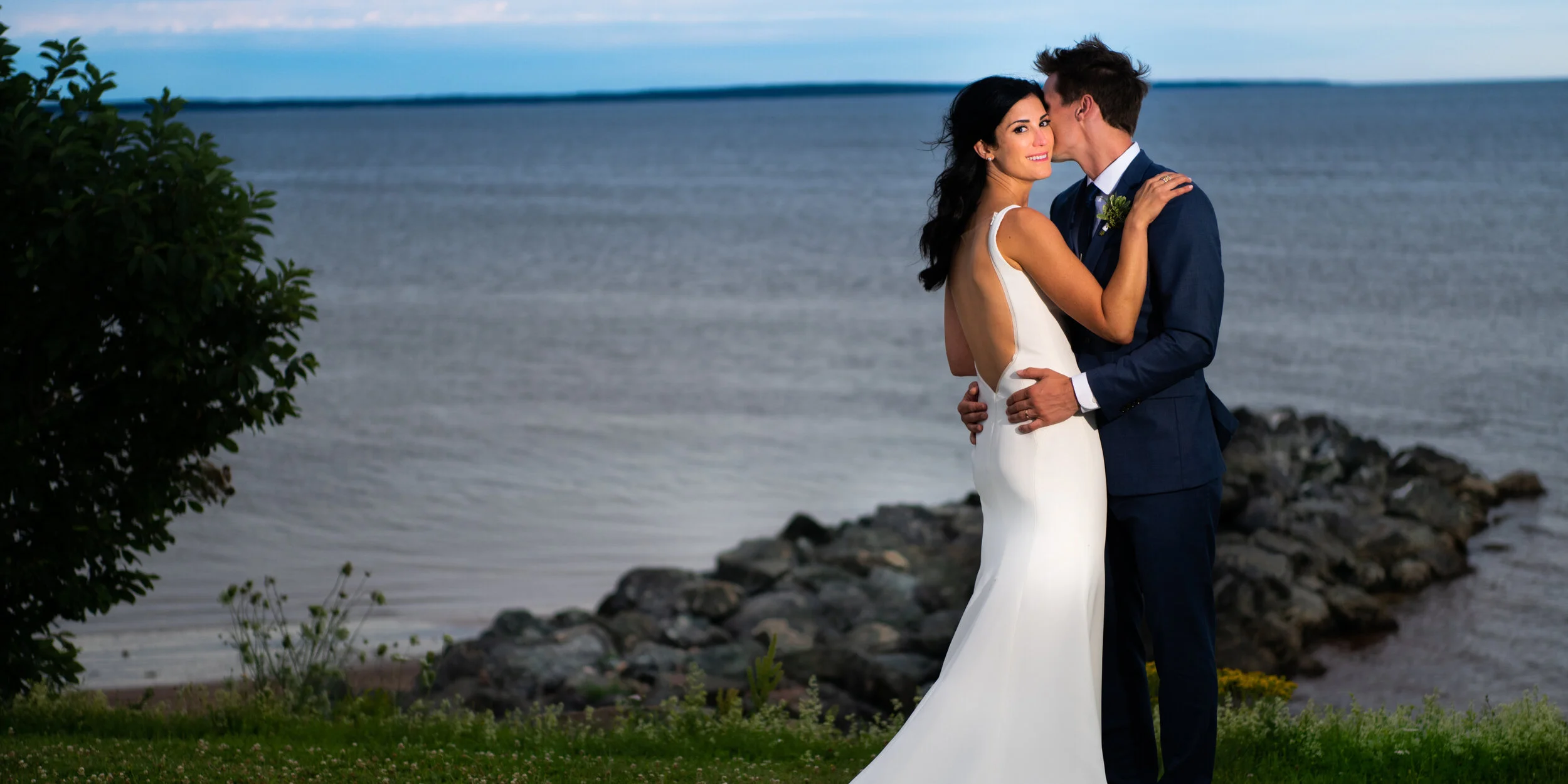 A couple dressed in wedding attire embracing by the water's edge, with a rocky shoreline and greenery in the foreground and a cloudy sky in the background.