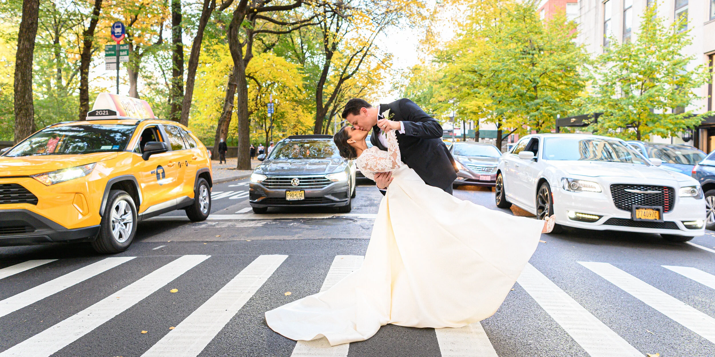 A bride and groom are dancing in the middle of a city street with cars parked and moving nearby. The bride is wearing a white wedding dress with lace sleeves, and the groom is in a black tuxedo. The street is lined with trees with green and yellow le
