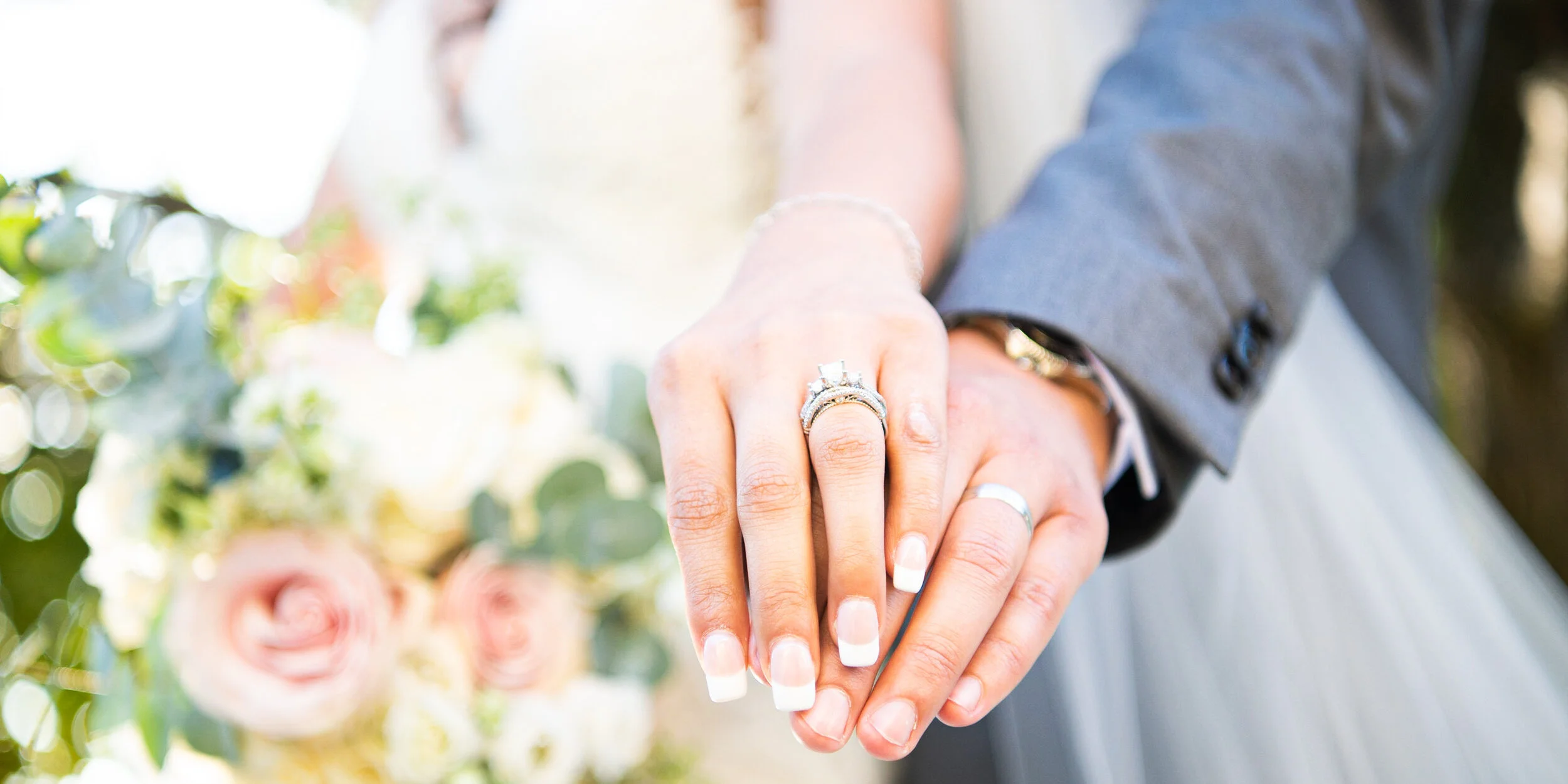 Close-up of a bride and groom holding hands, showing wedding rings, with blurred flowers in the background.