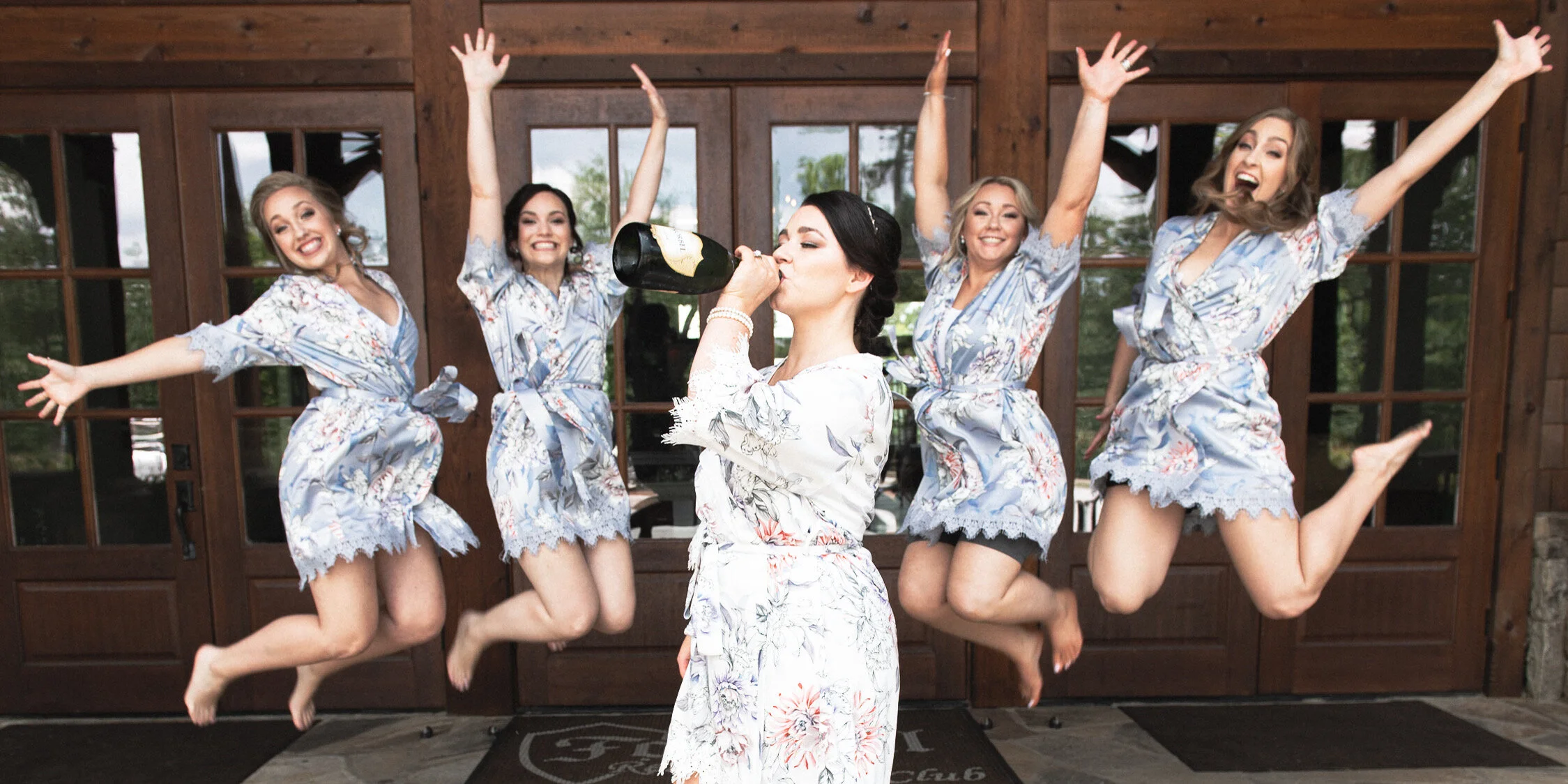 Group of five women in matching floral pajamas celebrating, with one woman drinking champagne while the others jump joyfully in front of a wooden door.