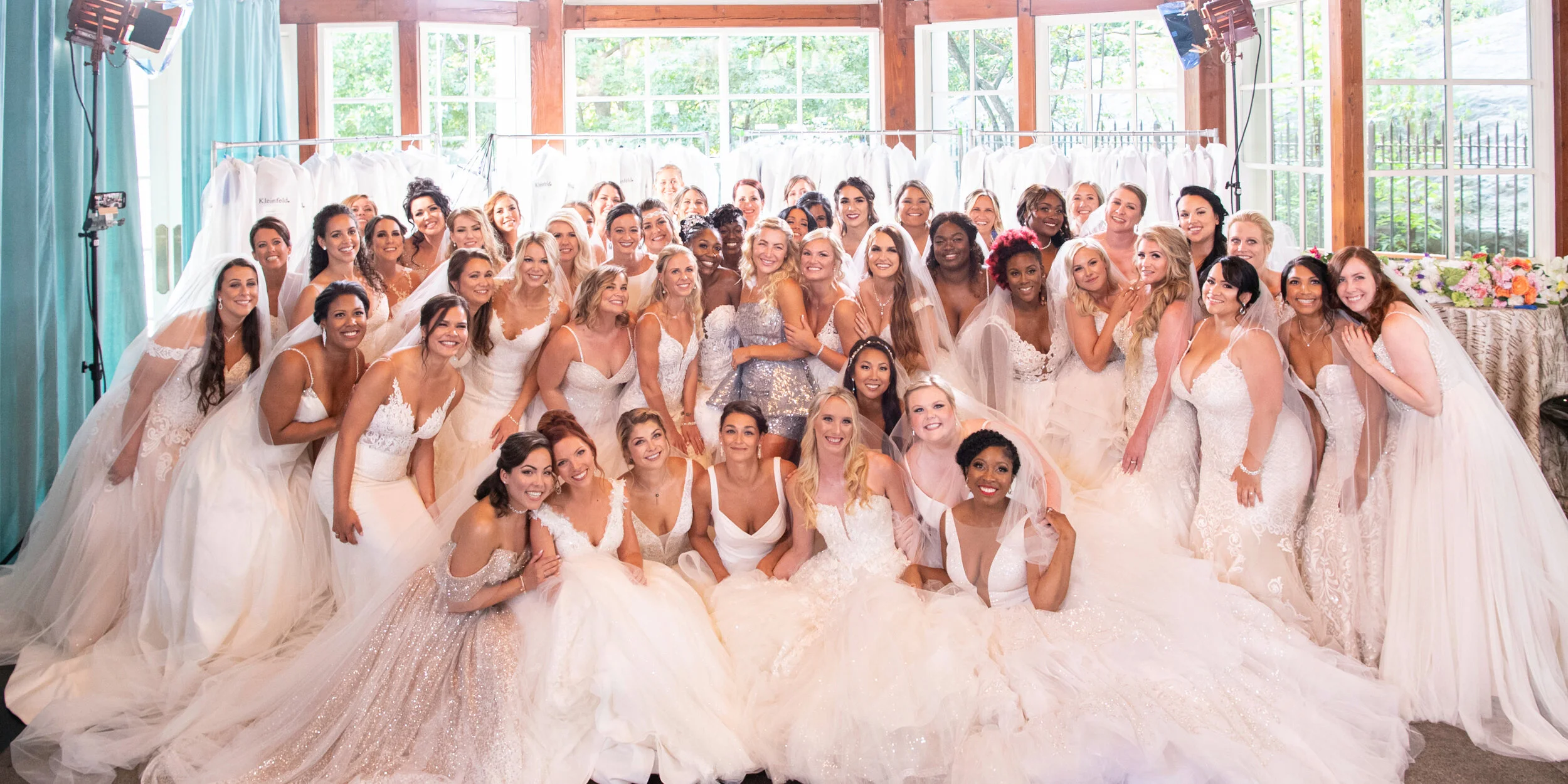 Group of women in wedding dresses posing in a brightly lit room with large windows and floral and clothing displays in the background.
