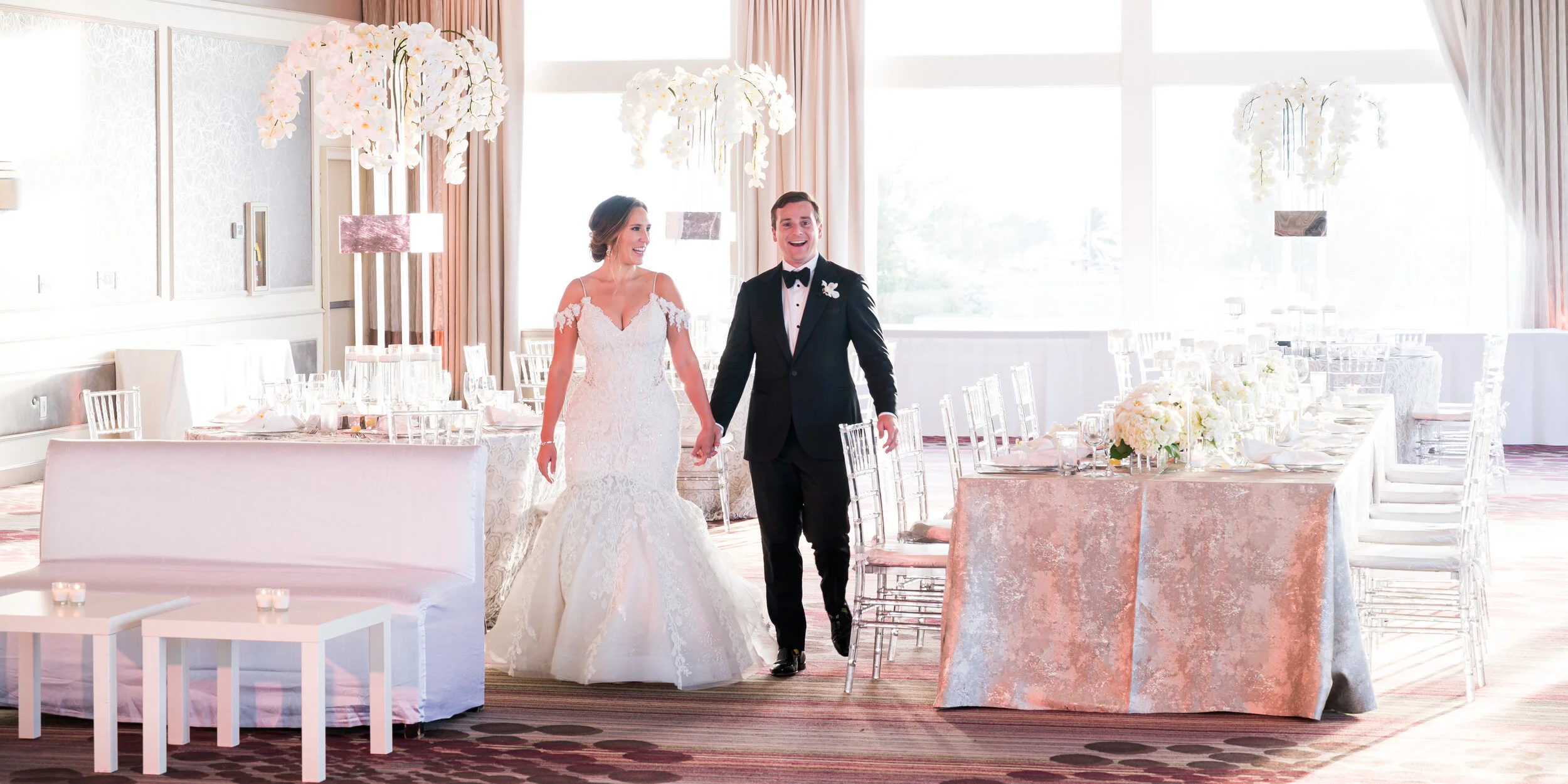 A bride and groom walking hand in hand at their wedding reception, decorated with white flowers and elegant table settings.