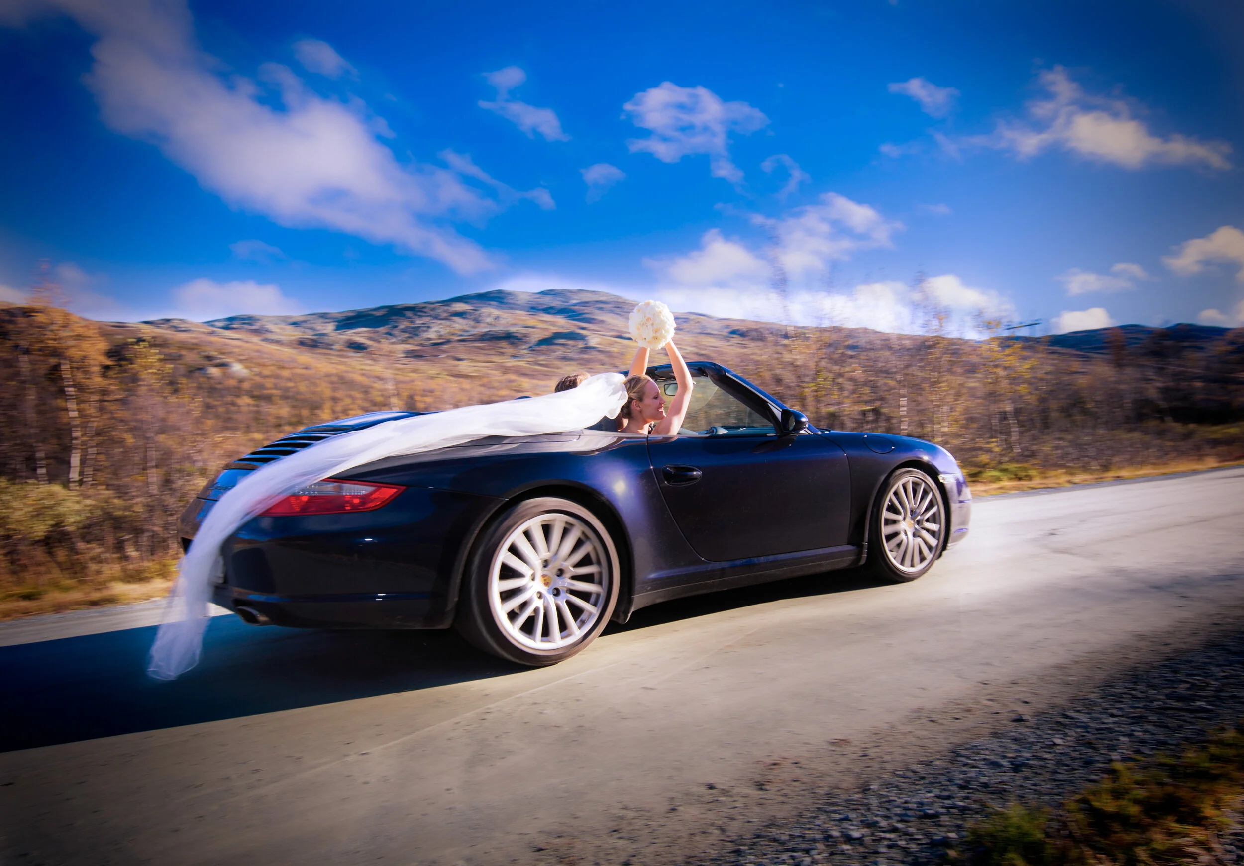 A woman in a wedding dress riding in a black convertible car on a road with mountains and blue sky in the background.