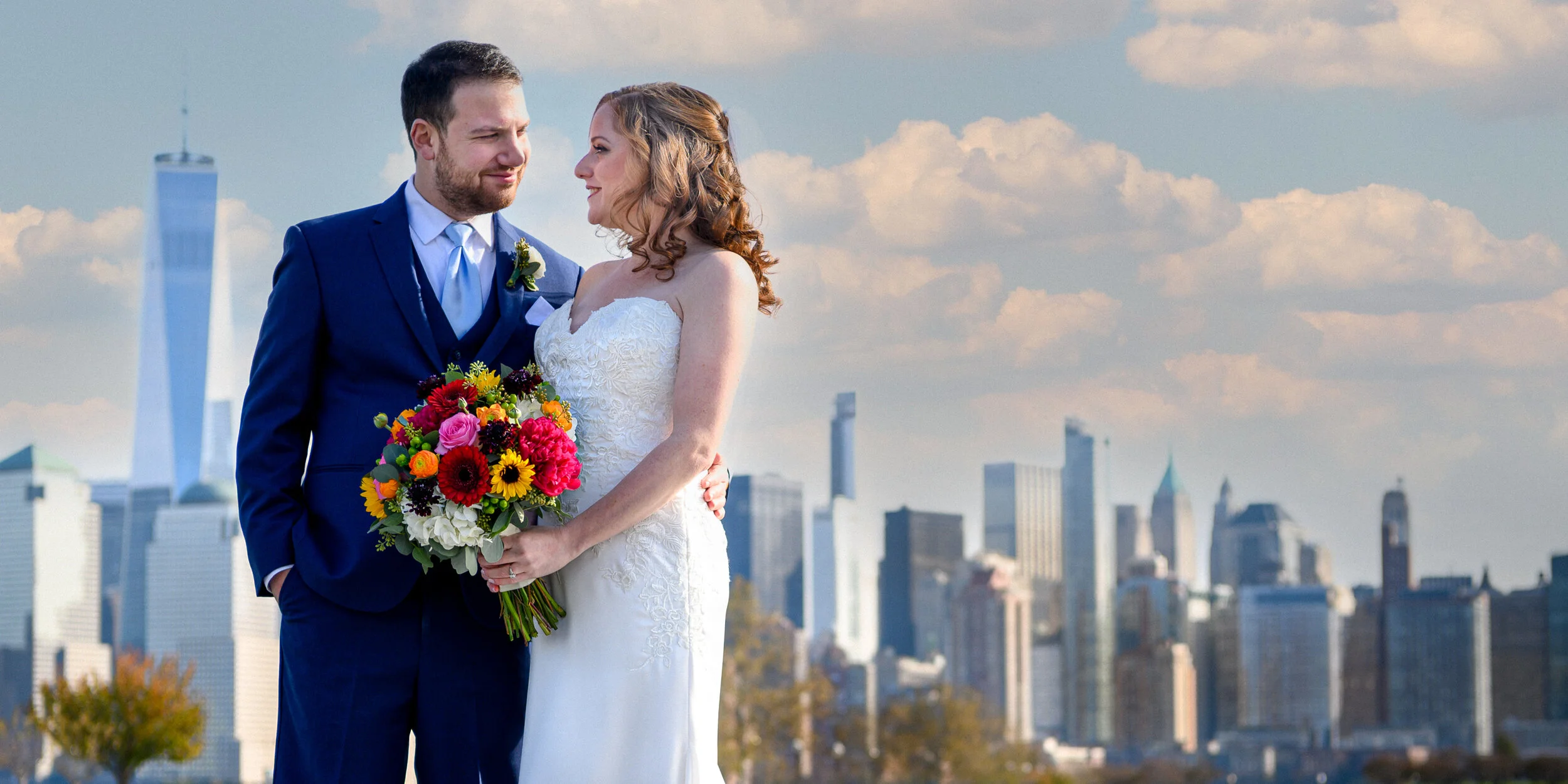 A bride and groom standing close together with New York City skyline in the background. The bride is holding a colorful bouquet of flowers and the groom is dressed in a blue suit.