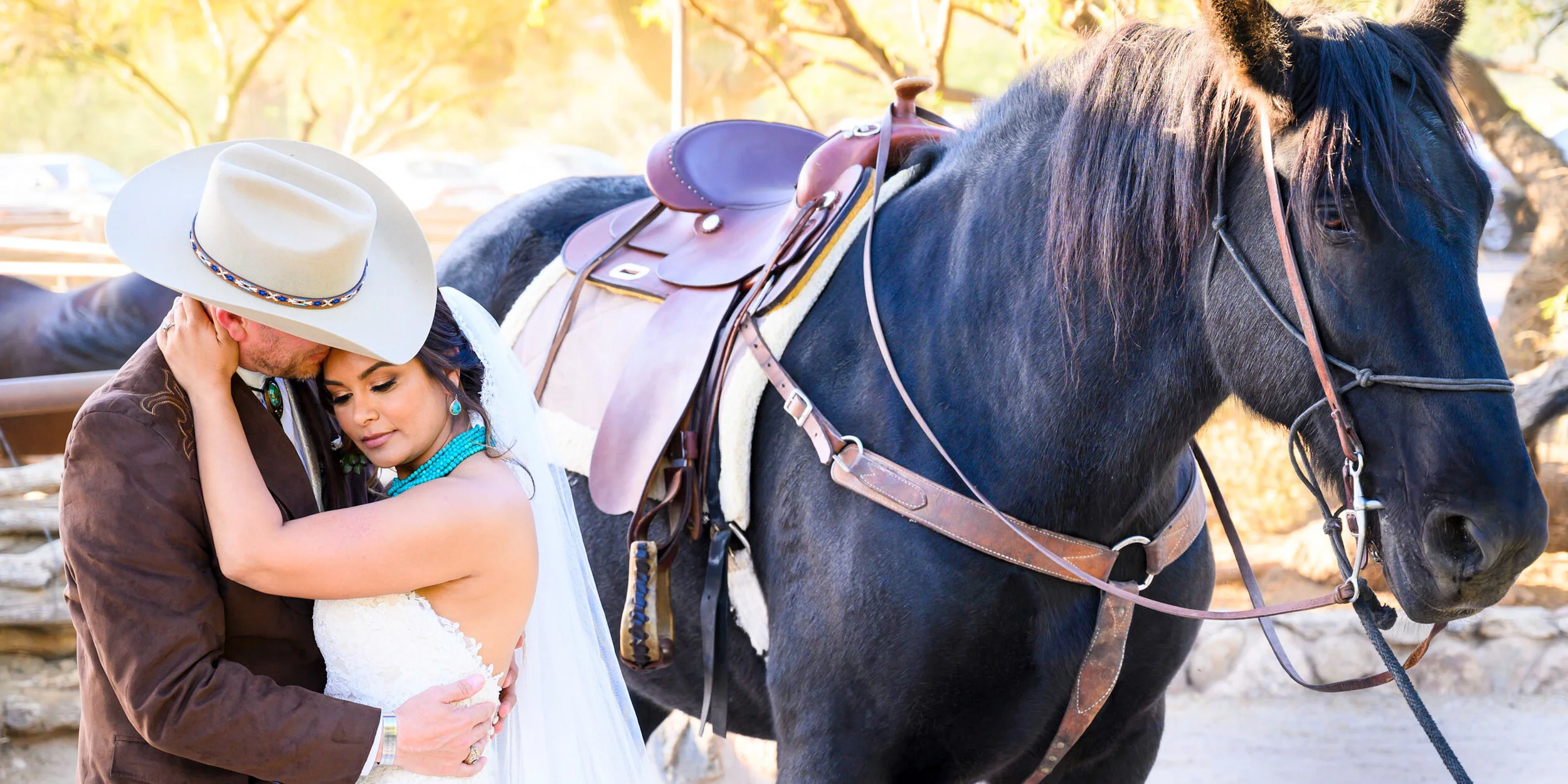 A man, woman dressed in wedding attire, hugging and looking at each other, standing next to a black horse with a saddle, outdoors during daytime with trees and cars in the background.