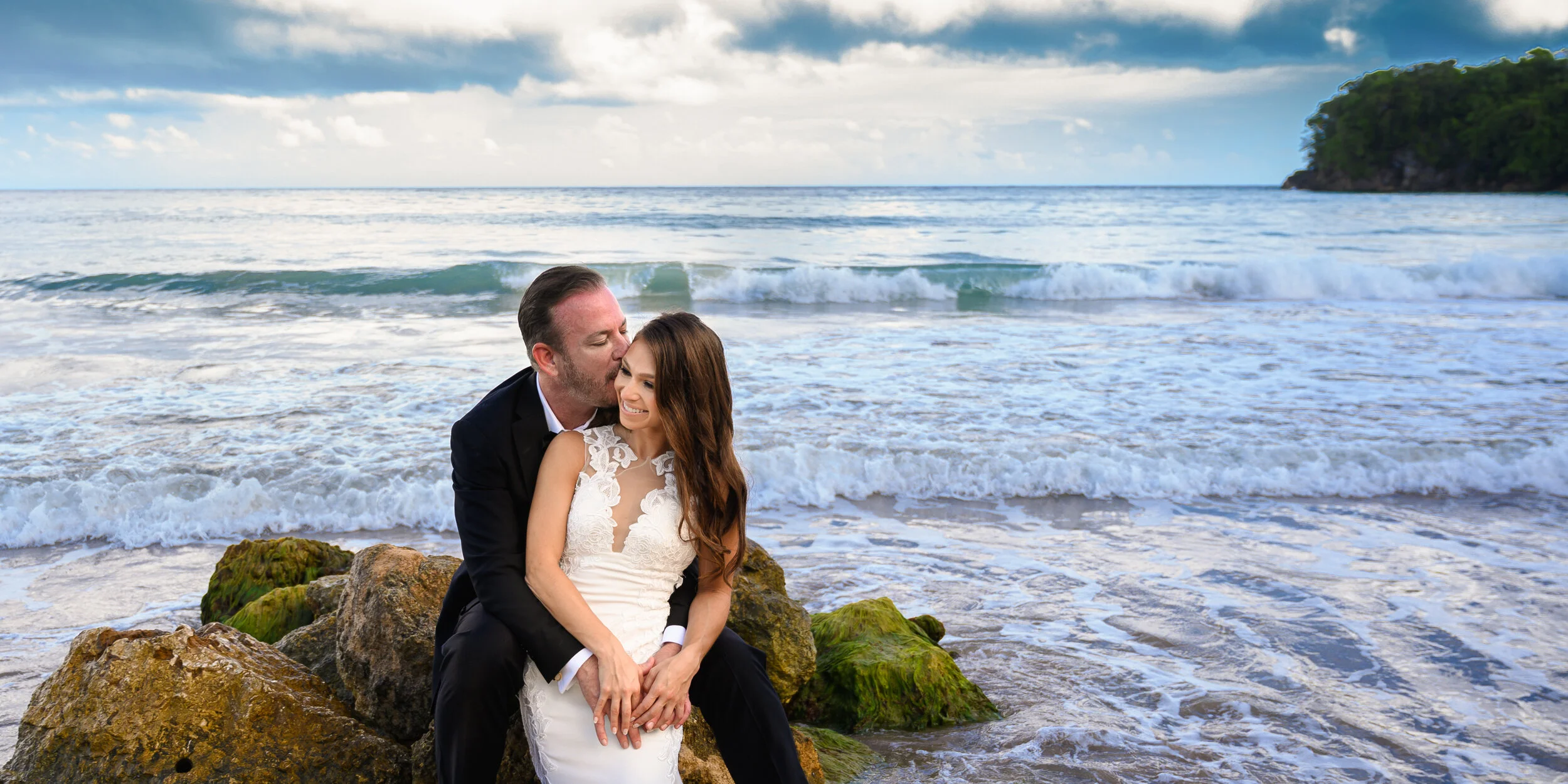 A couple sitting on rocks at the beach, with the man kissing the woman on the cheek. The woman is wearing a white lace dress, and the man is in a black suit. Waves are crashing on the shore, with a cloudy sky and a small green island in the backgroun