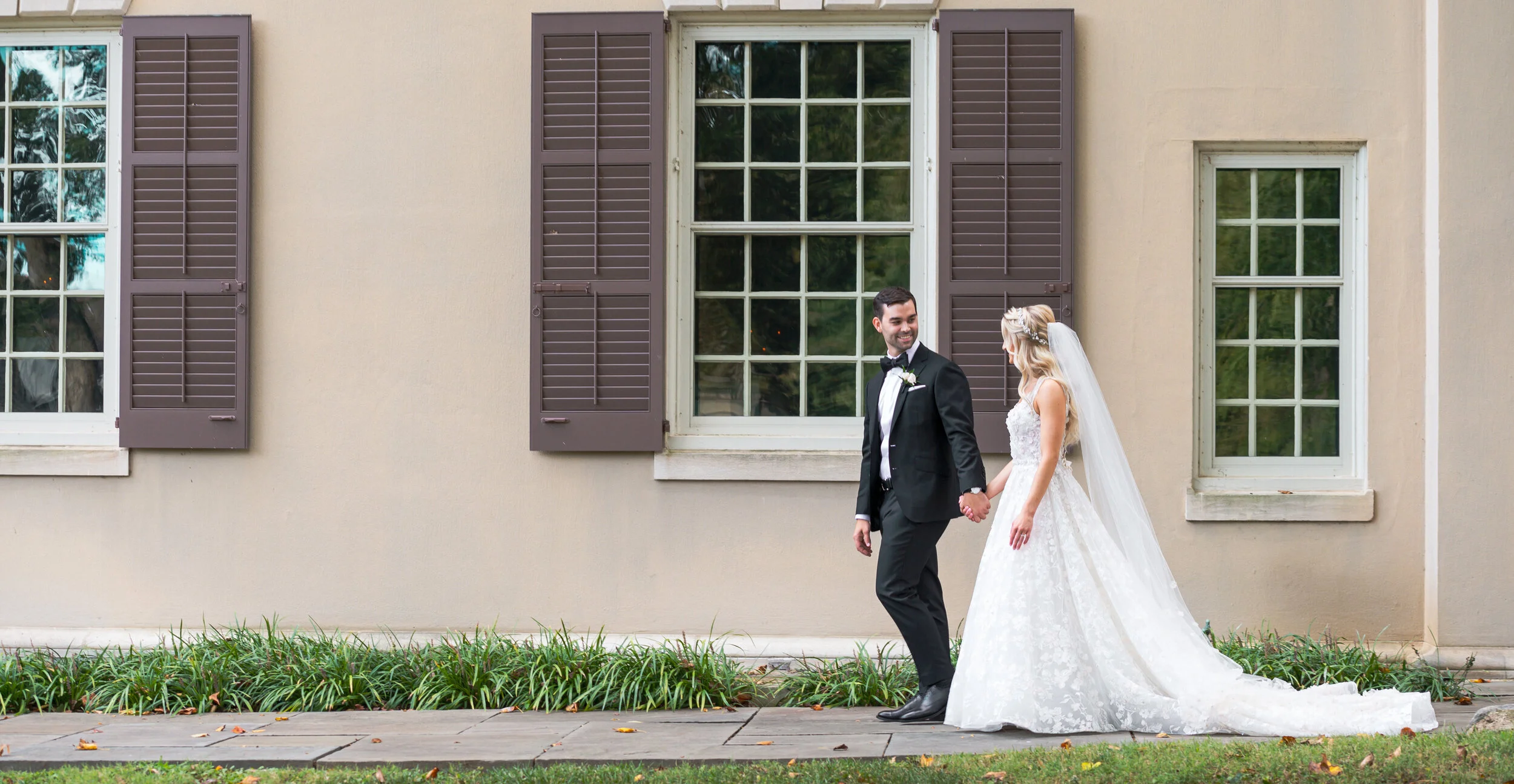 A newlywed couple walking hand in hand outside a building with beige walls and three windows with shutters. The groom is in a tuxedo, and the bride is in a white wedding dress with a long train and veil.
