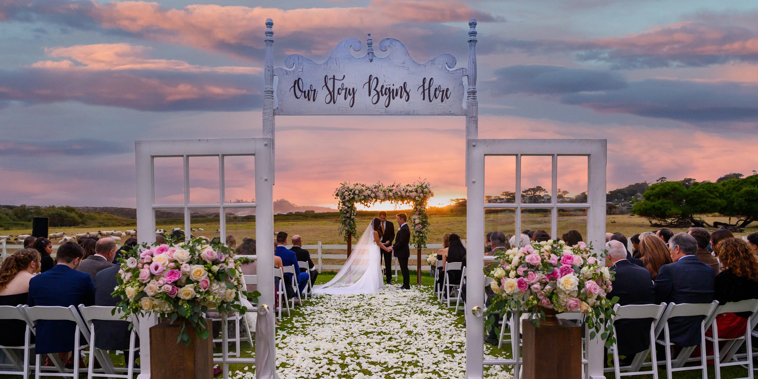 Couple getting married under an arch decorated with flowers during sunset, with guests seated and a scenic rural background with grazing sheep and trees.