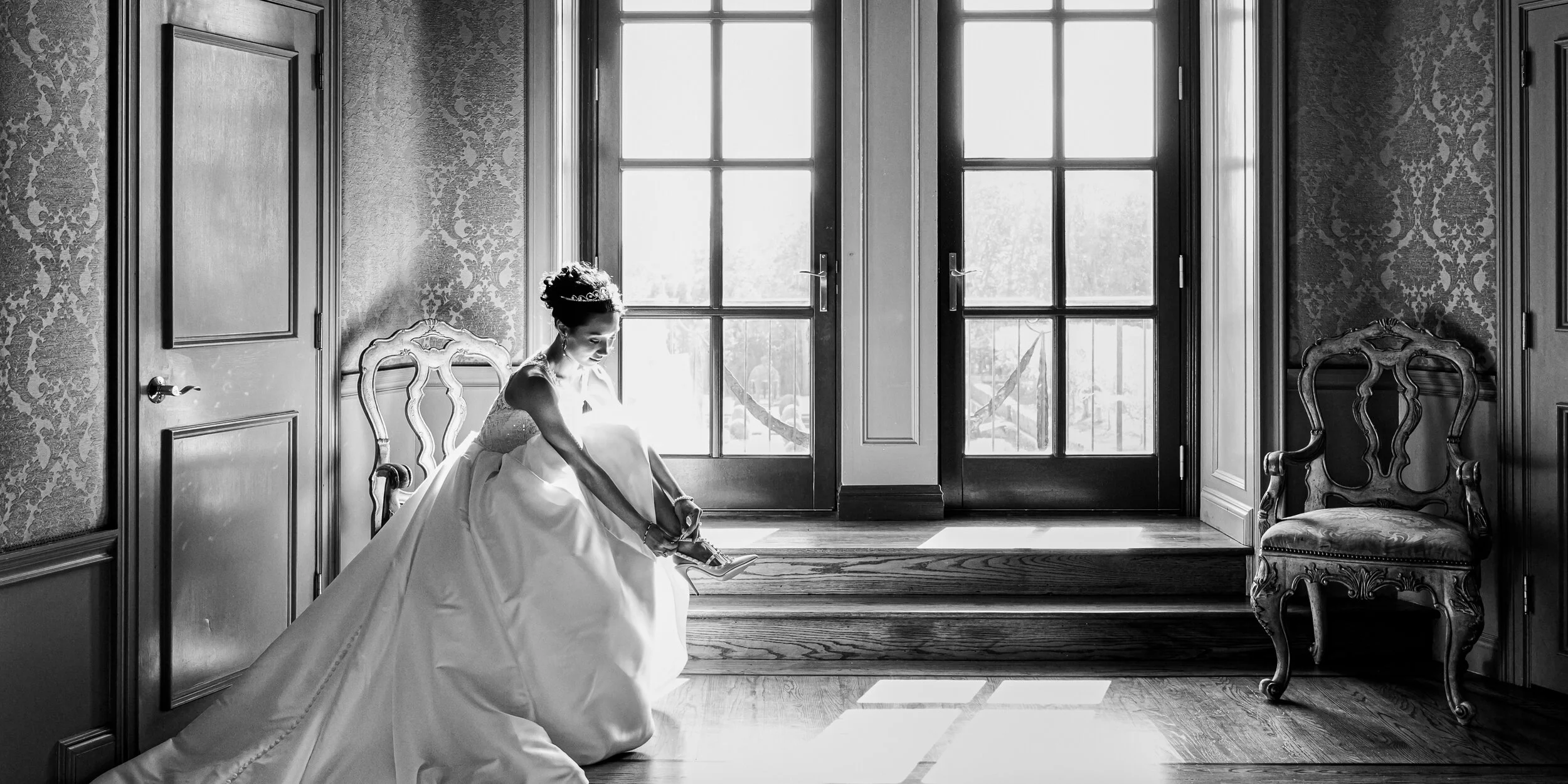 A black-and-white photo of a bride in a wedding gown sitting and putting on her high heel shoe in a bright room with large glass doors, ornate chairs, and patterned wallpaper.