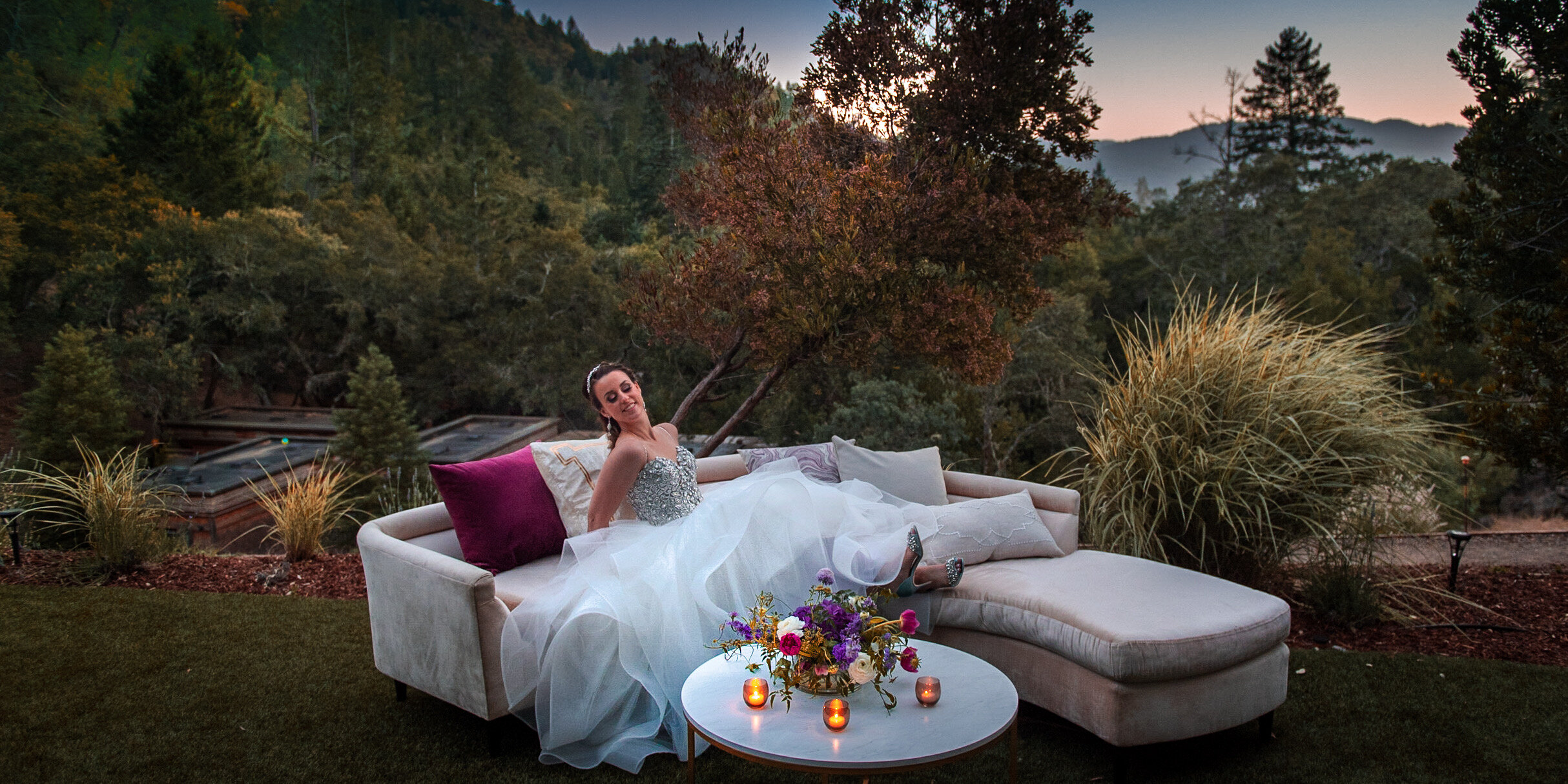 A woman in a white and silver dress is sitting on a light-colored outdoor sofa with purple and beige pillows, surrounded by trees and plants at dusk. There is a small table with candles and a bouquet of colorful flowers in front of her.