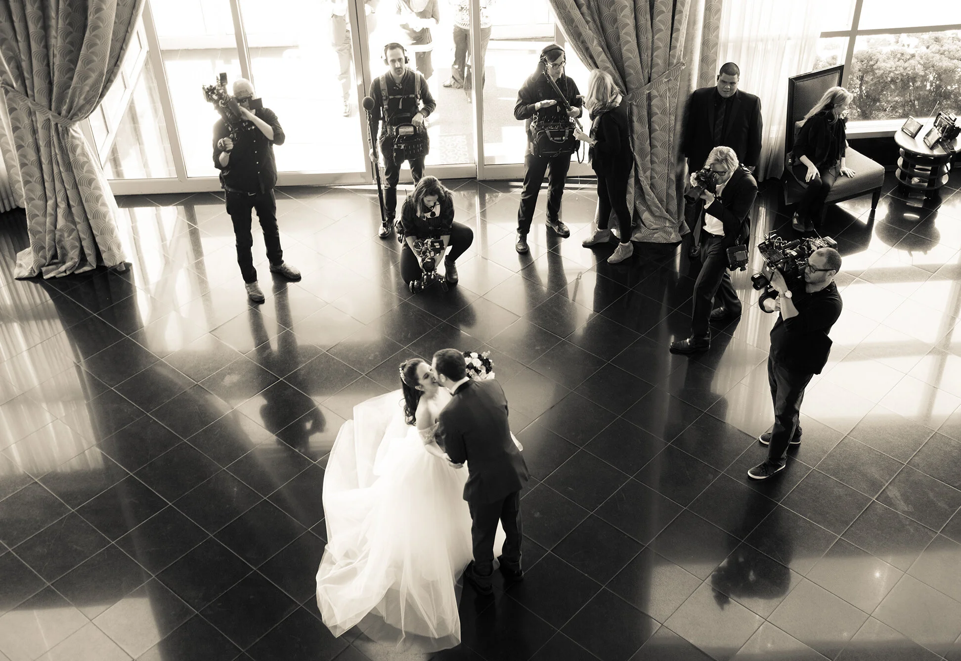 A wedding ceremony with the bride and groom kissing in front of a flower arrangement, surrounded by photographers and videographers capturing the moment in a spacious hall with large windows and curtains.