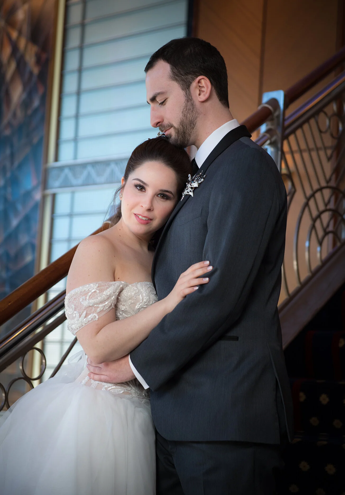 A bride and groom on a staircase, embracing affectionately, with the bride resting her head on the groom's forehead.