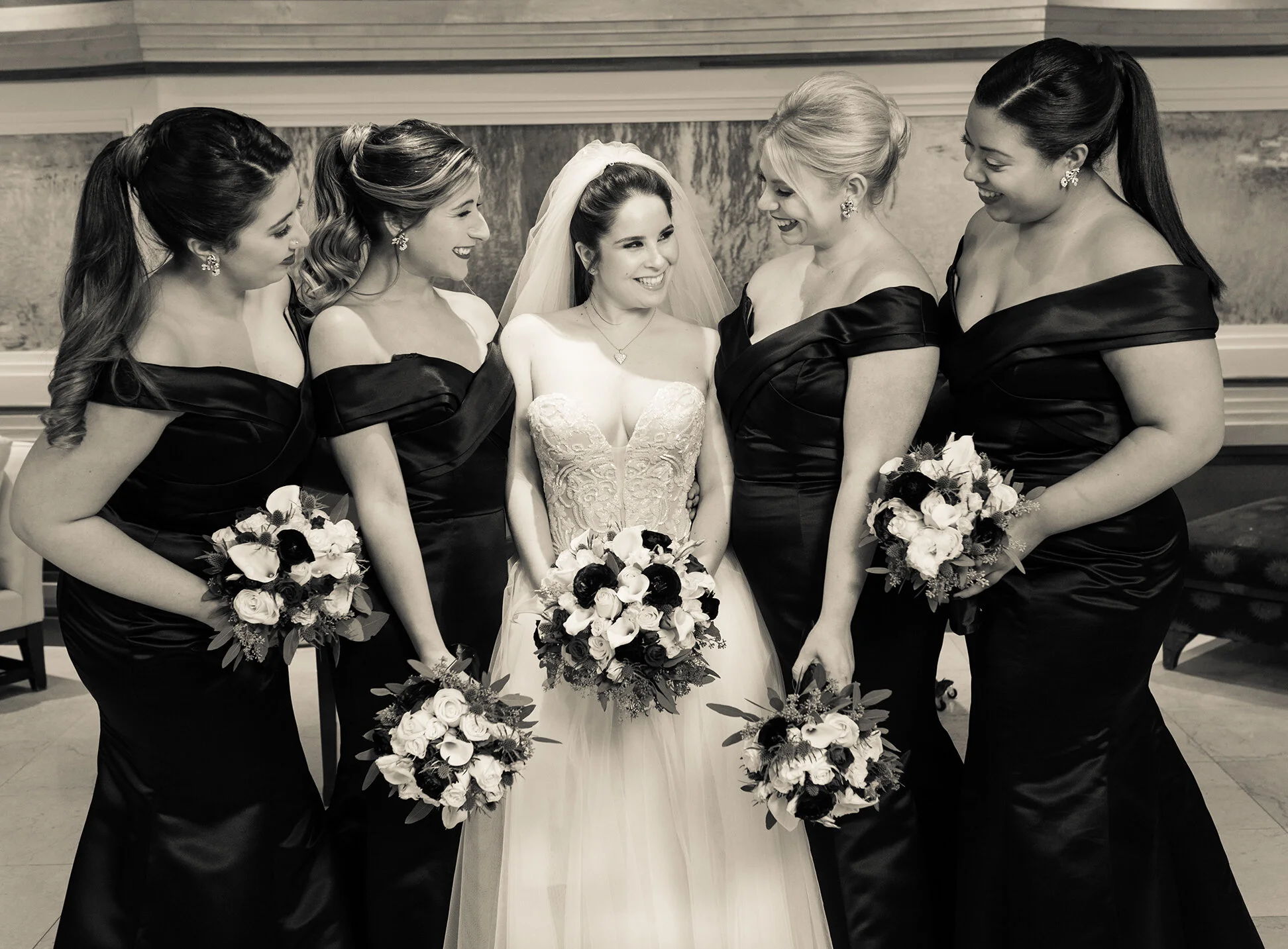 A bride in a wedding dress surrounded by five women in black dresses, holding bouquets of flowers, at a wedding celebration.