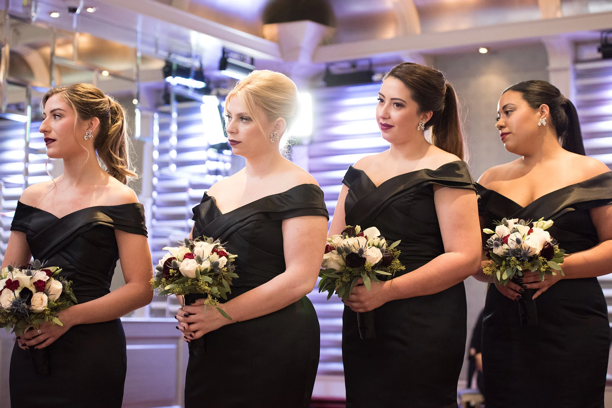 Four women dressed in elegant black off-the-shoulder dresses stand in a line holding bouquets of white, black, and dark red flowers during what appears to be a formal event or wedding.