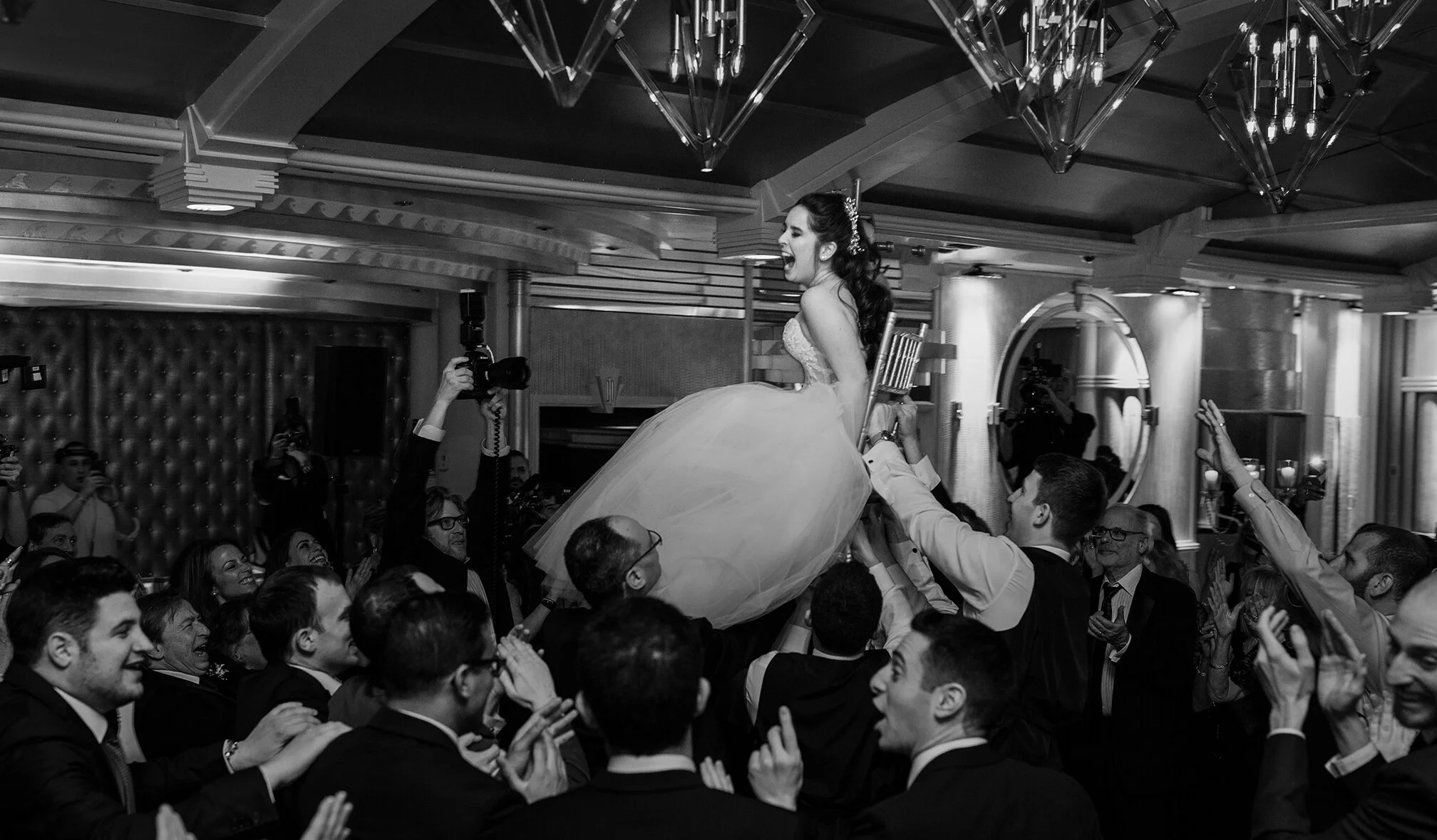 A bride being lifted on a chair during her wedding celebration, surrounded by guests clapping and smiling in an elegant, decorated indoor venue.