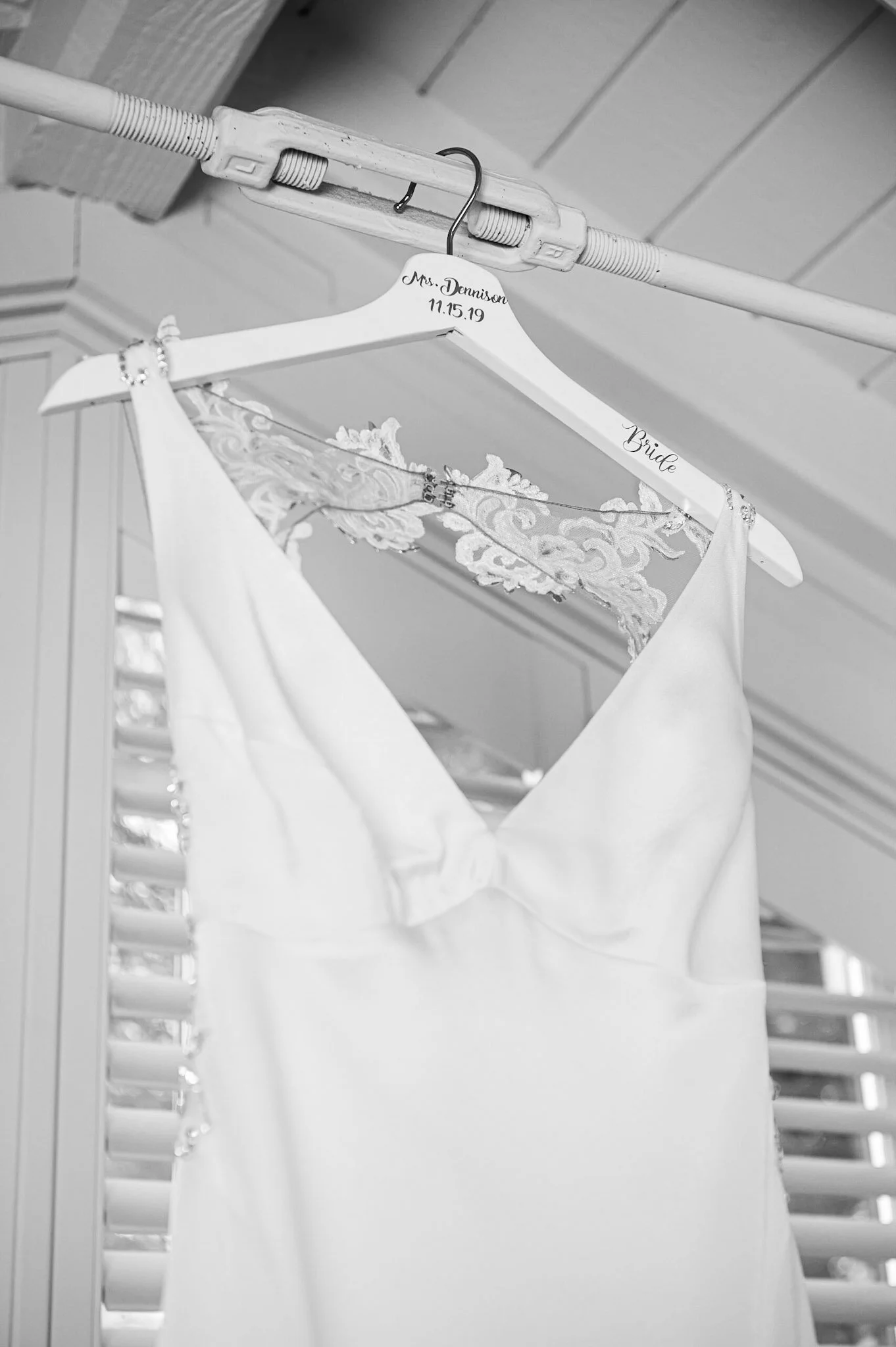 A white wedding dress hanging on a hanger labeled with 'Mrs. Denison' and the date '11.15.19' on a decorative hanger that says 'Bride'. The dress is attached to a clothes rod and the background shows wooden paneling.