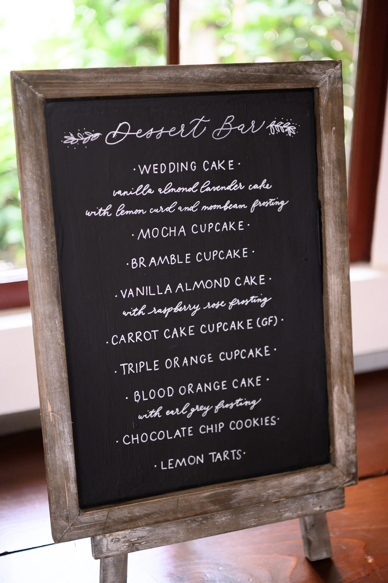 A black chalkboard with a wooden frame displaying a dessert bar menu, written in white cursive and print lettering, set on a wooden stand with a blurred green outdoor background.