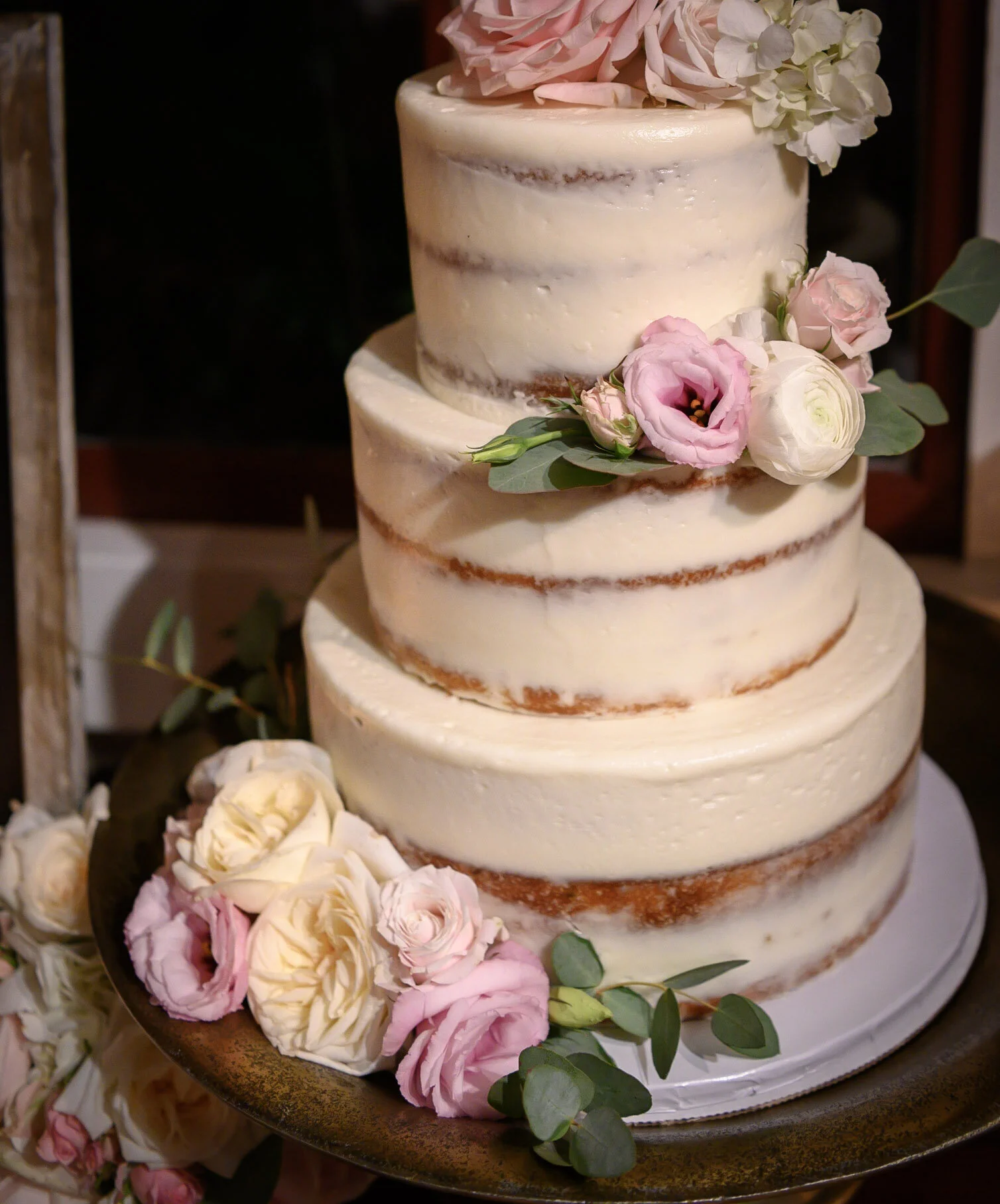 A three-tier wedding cake with a semi-naked frosting style decorated with pink and white roses and green leaves.