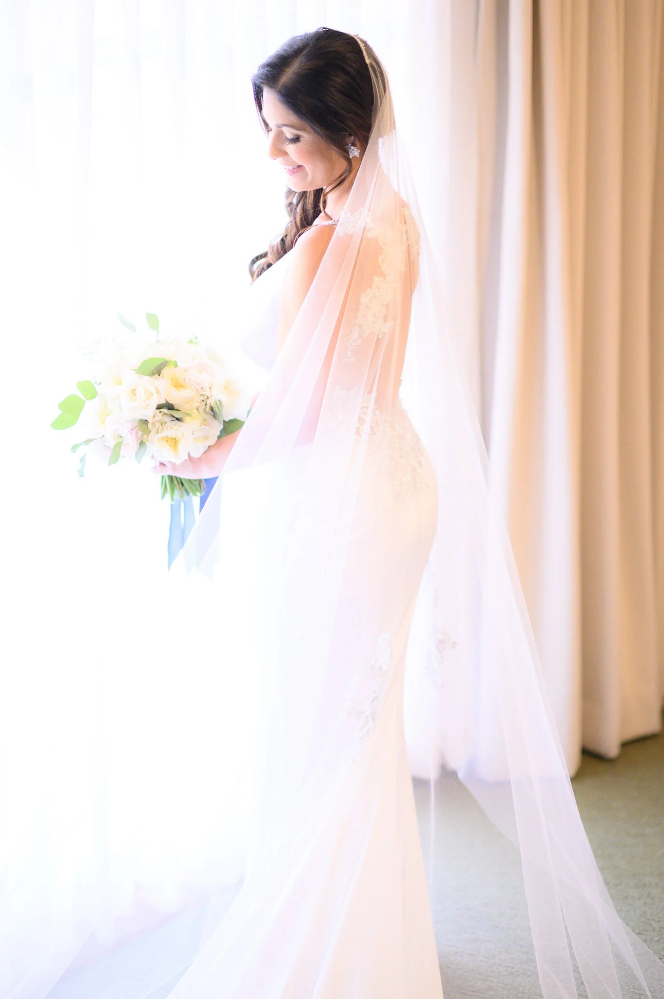 A woman in a wedding dress holding a bouquet of white roses, standing near a window with beige curtains, smiling with closed eyes.