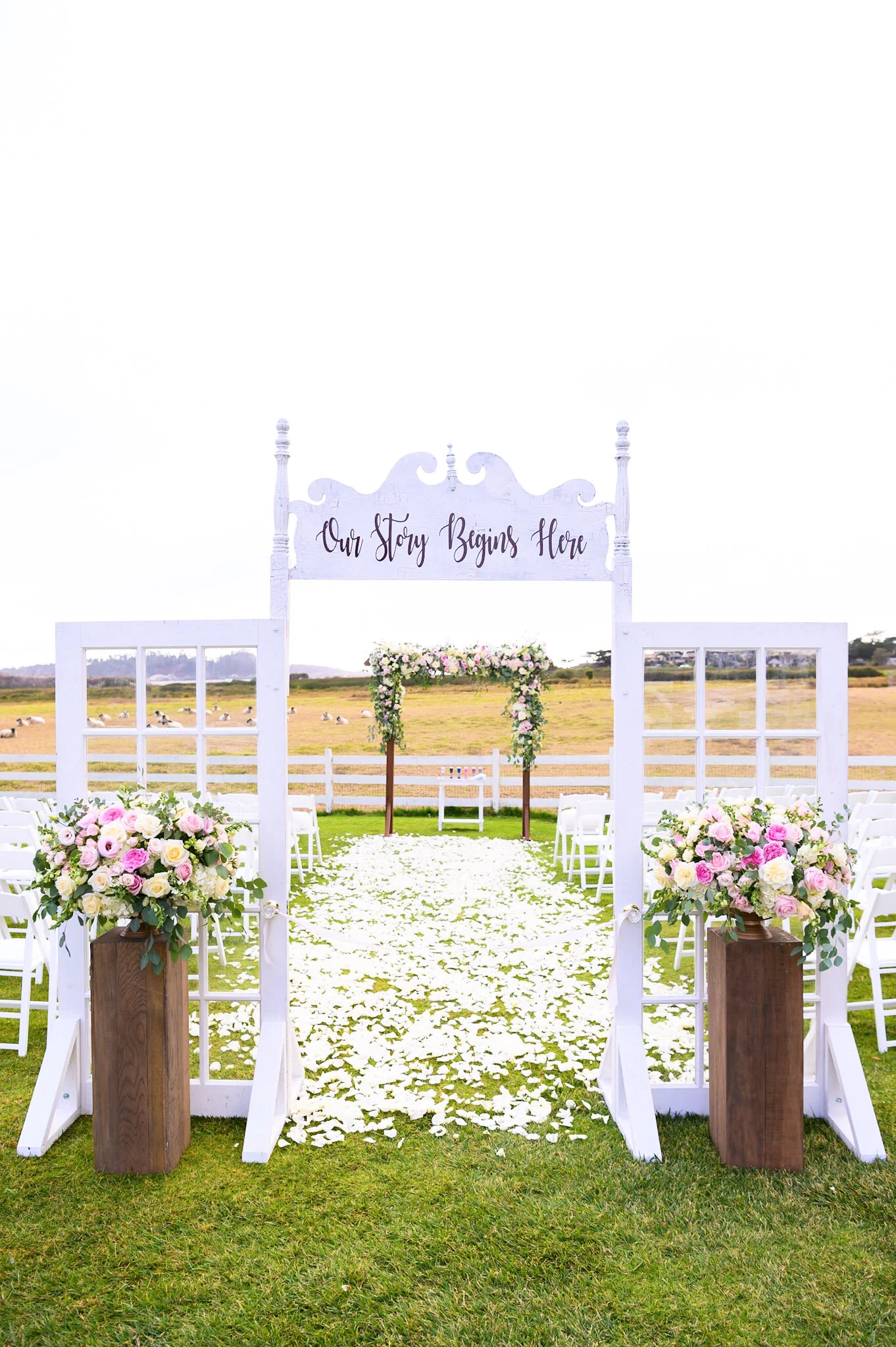 Outdoor wedding ceremony setup with white wooden arch decorated with pink and white flowers, white chairs, and a flower-covered aisle on a green lawn with a field and distant houses in the background.