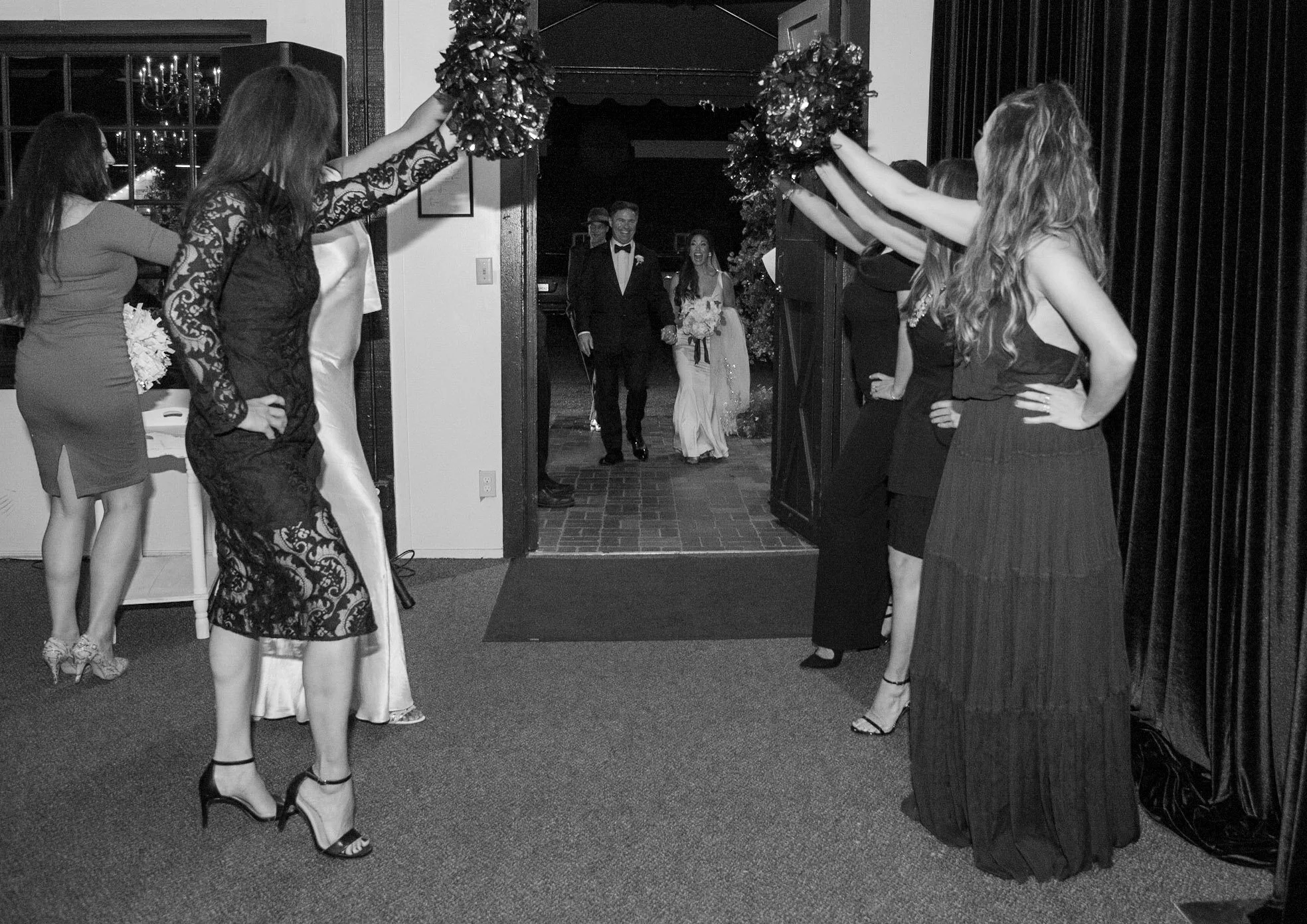 Guests at a wedding reception enter through an open door decorated with greenery and flowers. A bride and groom are walking into the room, smiling and holding hands, while the bridal party stands nearby.