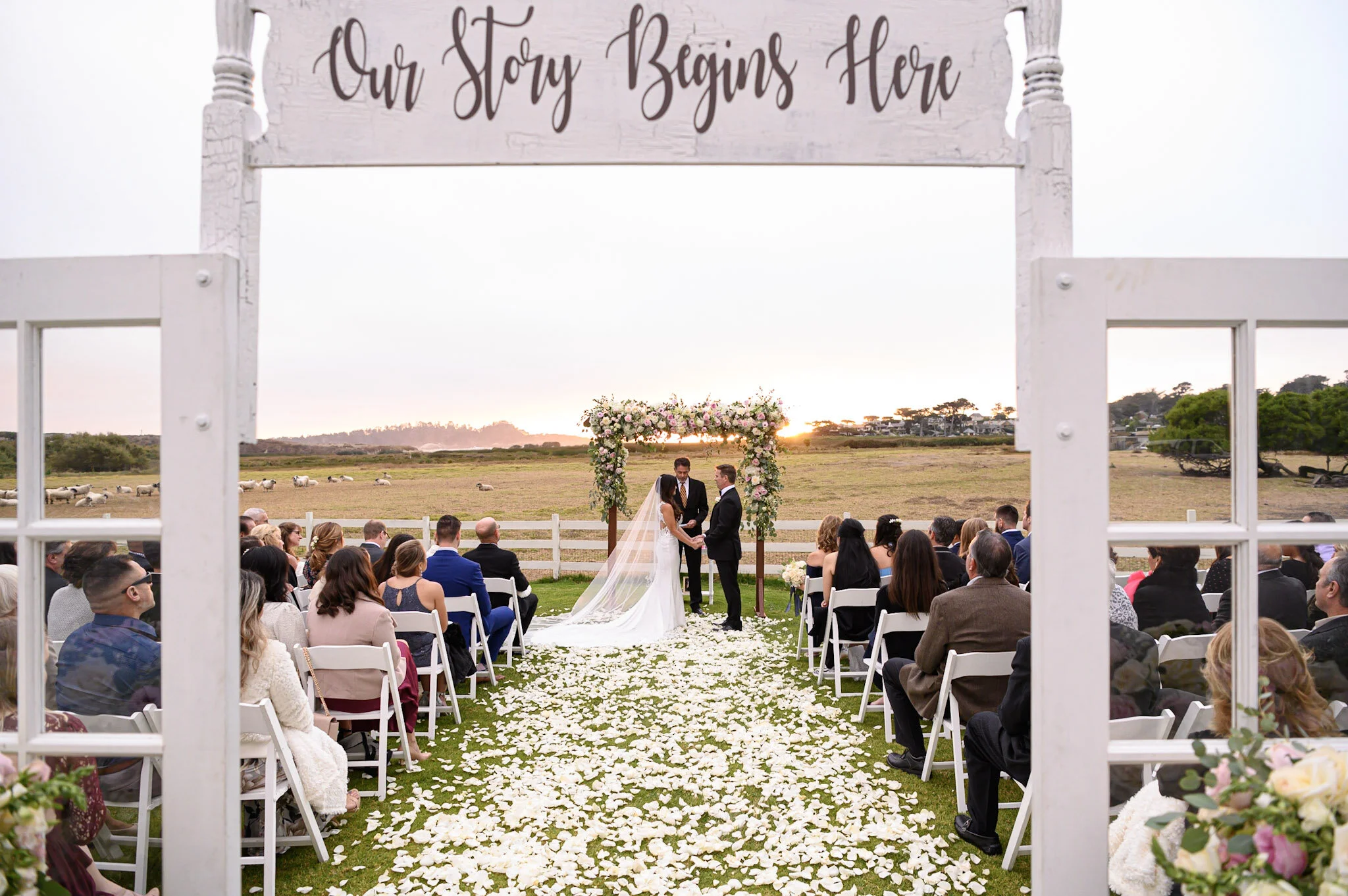 An outdoor wedding ceremony with a couple exchanging vows under a floral arch at sunset, with guests seated on white chairs on grass, rose petals on the aisle, and a scenic rural landscape in the background.