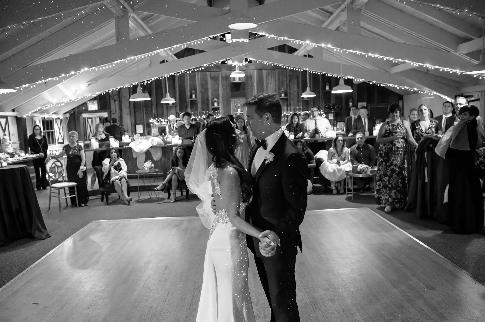 A bride and groom dance at their wedding reception in a rustic barn decorated with string lights and candles.
