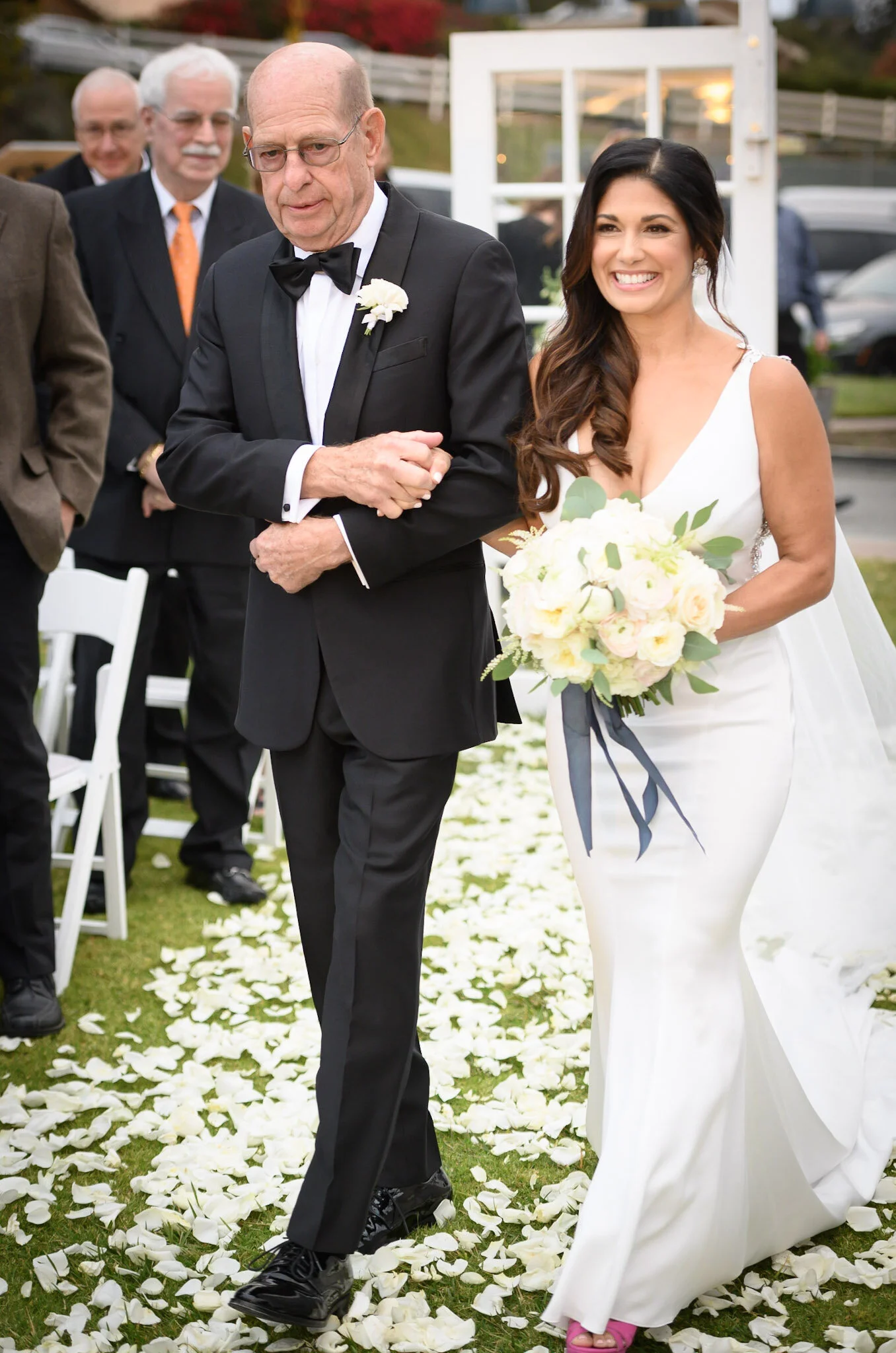 A bride in a white wedding dress carrying a bouquet of white and yellow roses walks down an outdoor aisle lined with white rose petals, escorted by an older man in a tuxedo, with three other men dressed in suits following behind.