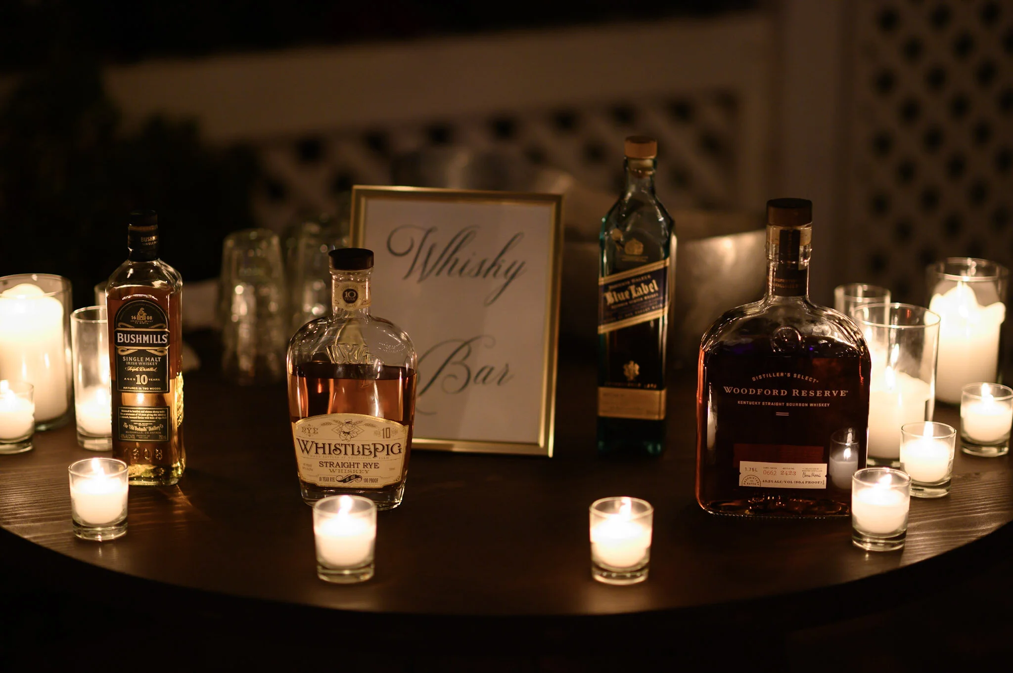 A display of whiskey bottles and candles on a table, with a card labeled 'Whisky Bar' in the background, creating a cozy ambiance.