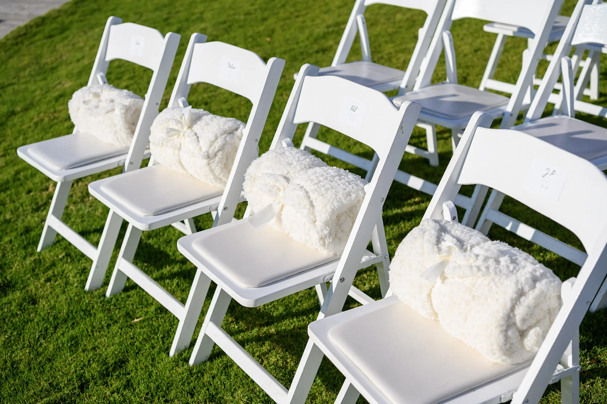 White folding chairs with fluffy white decorations tied to the backrests, arranged on a grassy outdoor area.