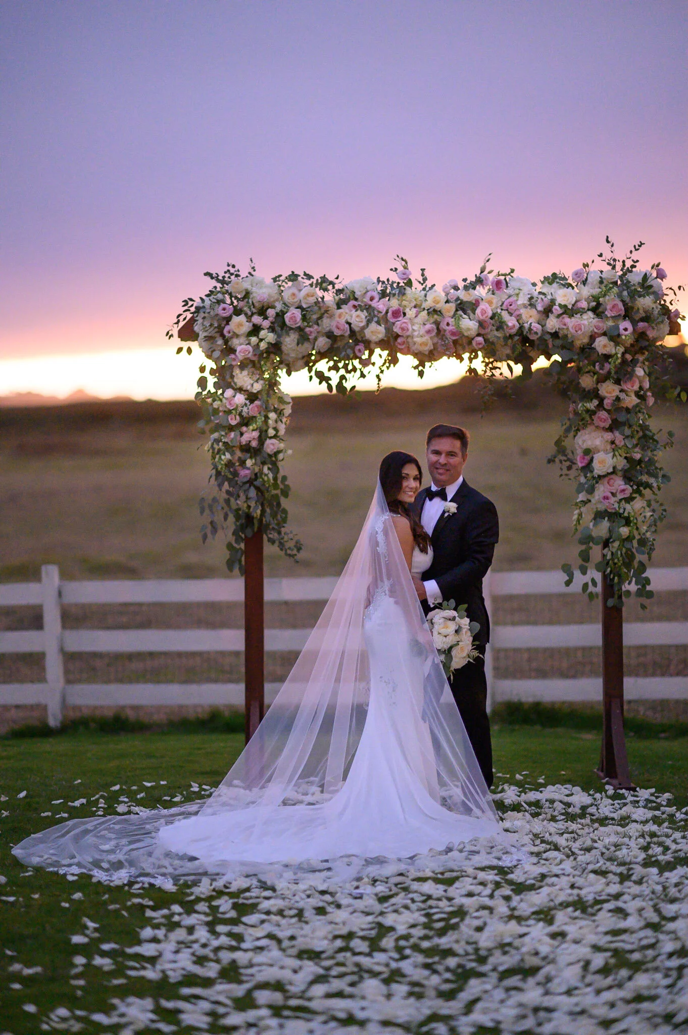 A bride and groom standing under a floral wedding arch during sunset at an outdoor wedding ceremony, with white flower petals scattered on the grass.