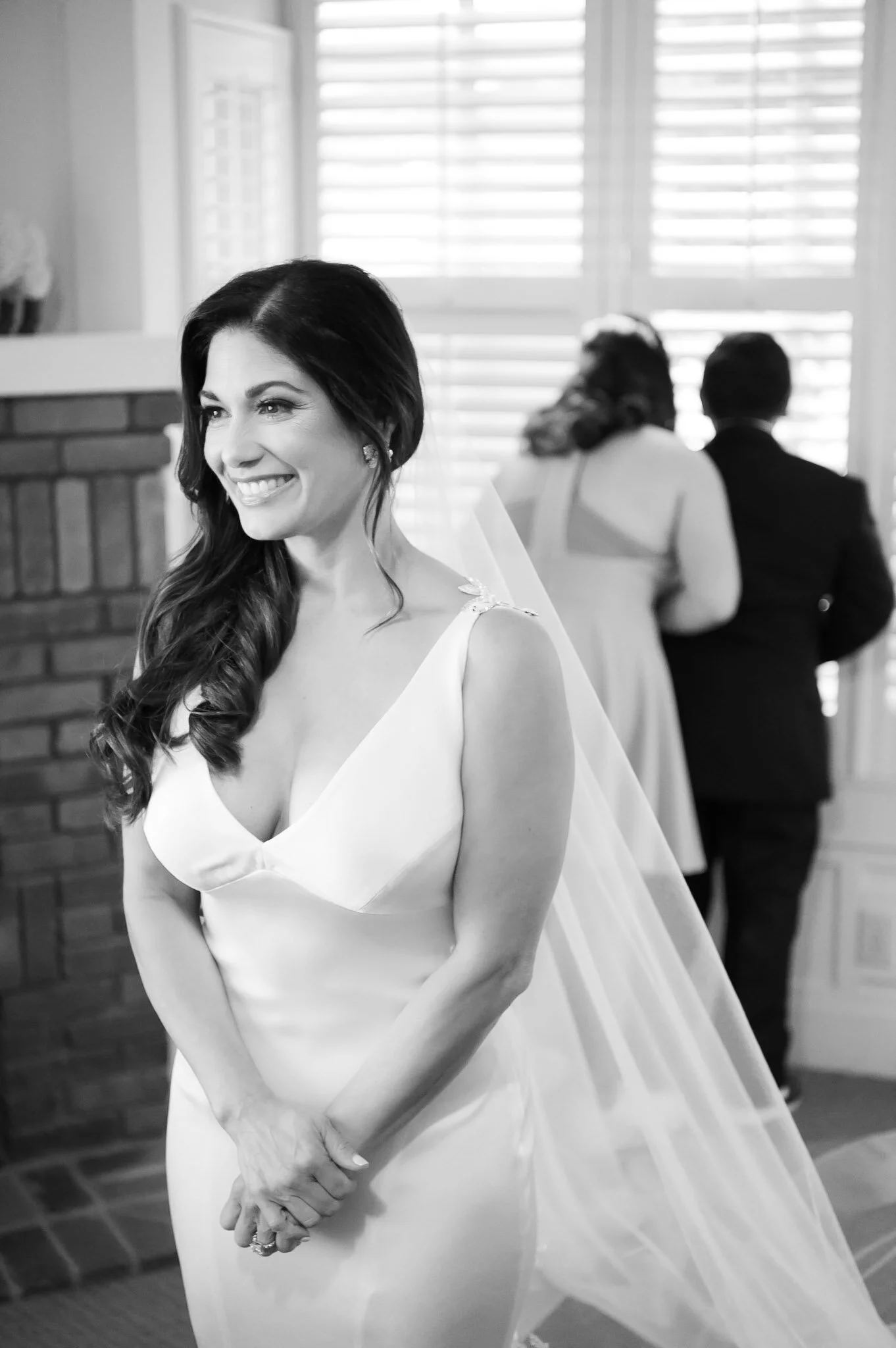 A smiling woman in a wedding dress standing with her hands clasped in front of her, with couple in formal attire in the background facing away, inside a room with large window blinds.