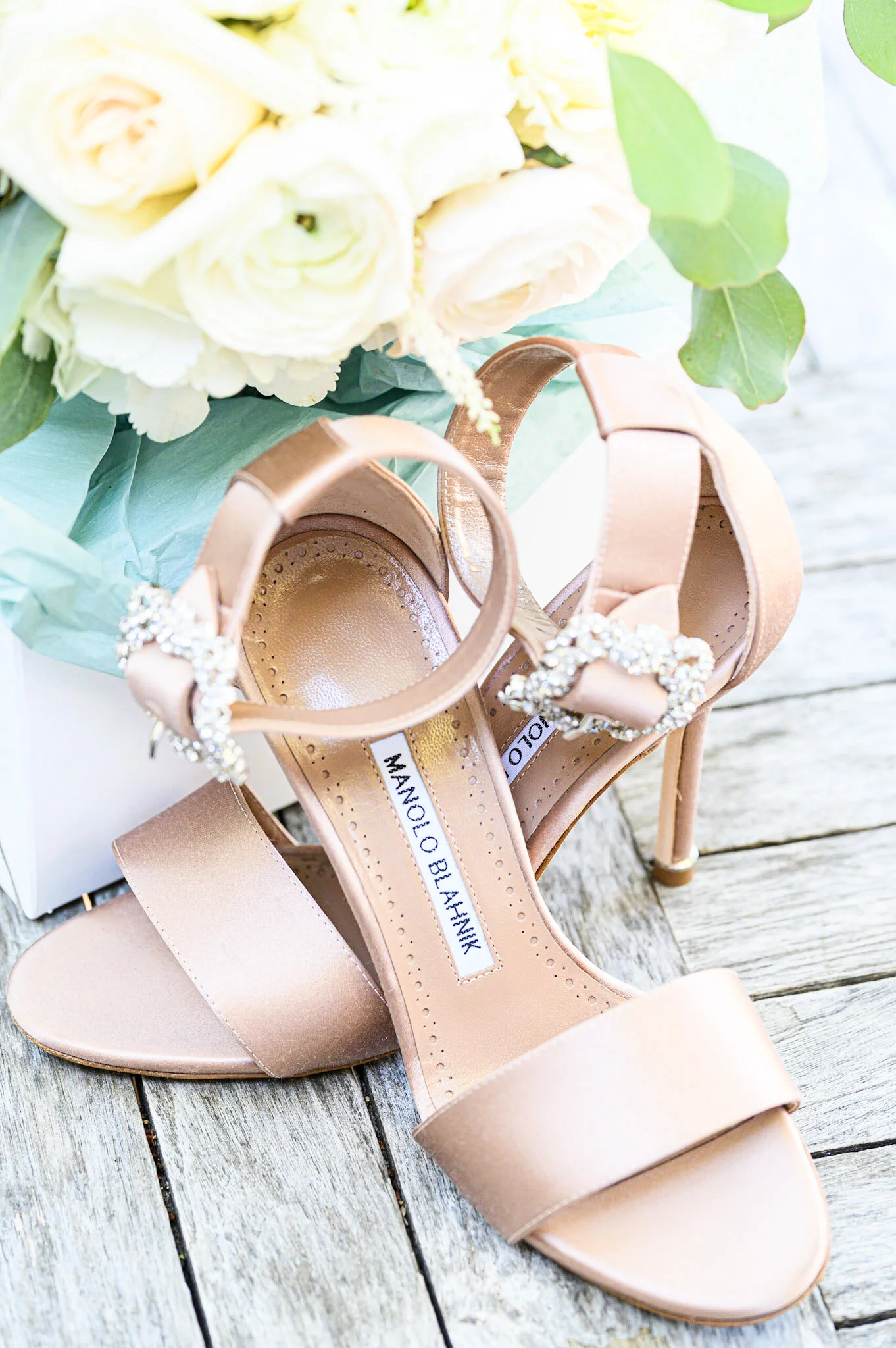 A pair of blush pink high-heeled sandals with rhinestone embellishments on the straps, placed on a wooden surface. A bouquet of white flowers with green leaves is partially visible in the background.
