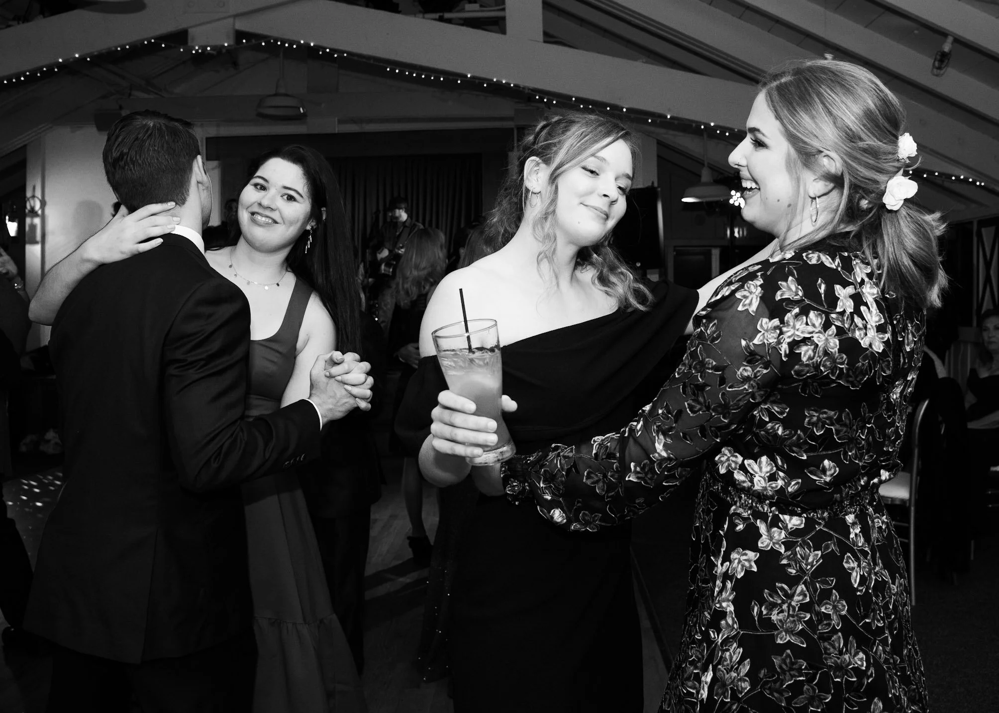 Women and a man dancing at a social event. The women are smiling and holding drinks, and the man is facing away from the camera.