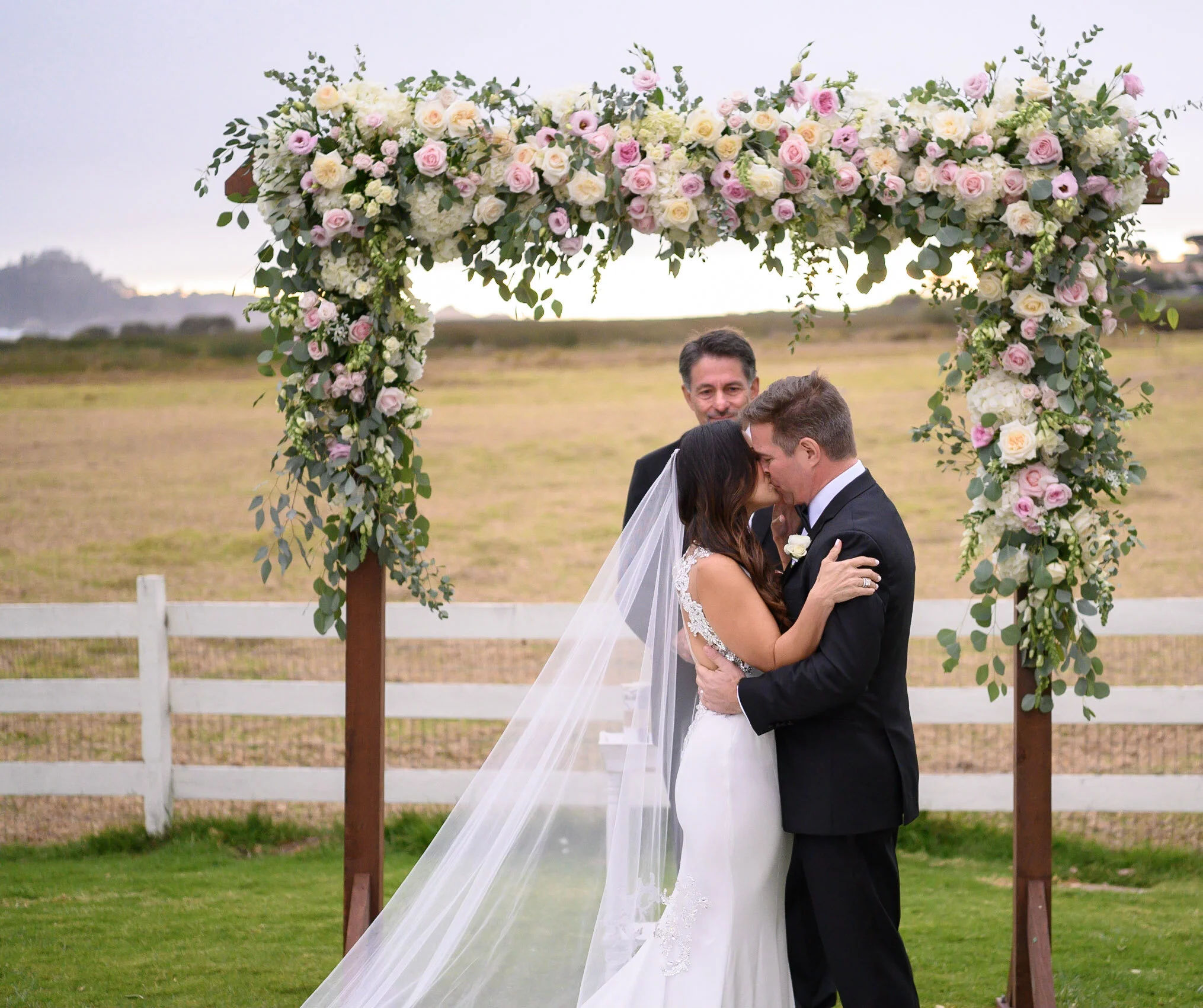 A wedding ceremony outdoors with a floral archway of pink and white roses and greens, where a bride and groom are kissing, with a smiling officiant in the background and a white fence and open field in the background.