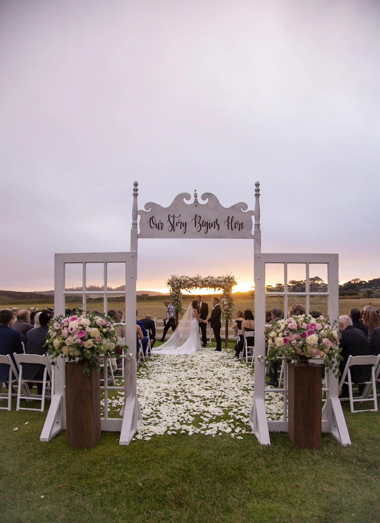 Outdoor wedding ceremony with a bride and groom exchanging vows under an arch decorated with flowers at sunset, guests seated on either side, white petals scattered on the aisle, and a sign reading "Our Story Begins Here" overhead.